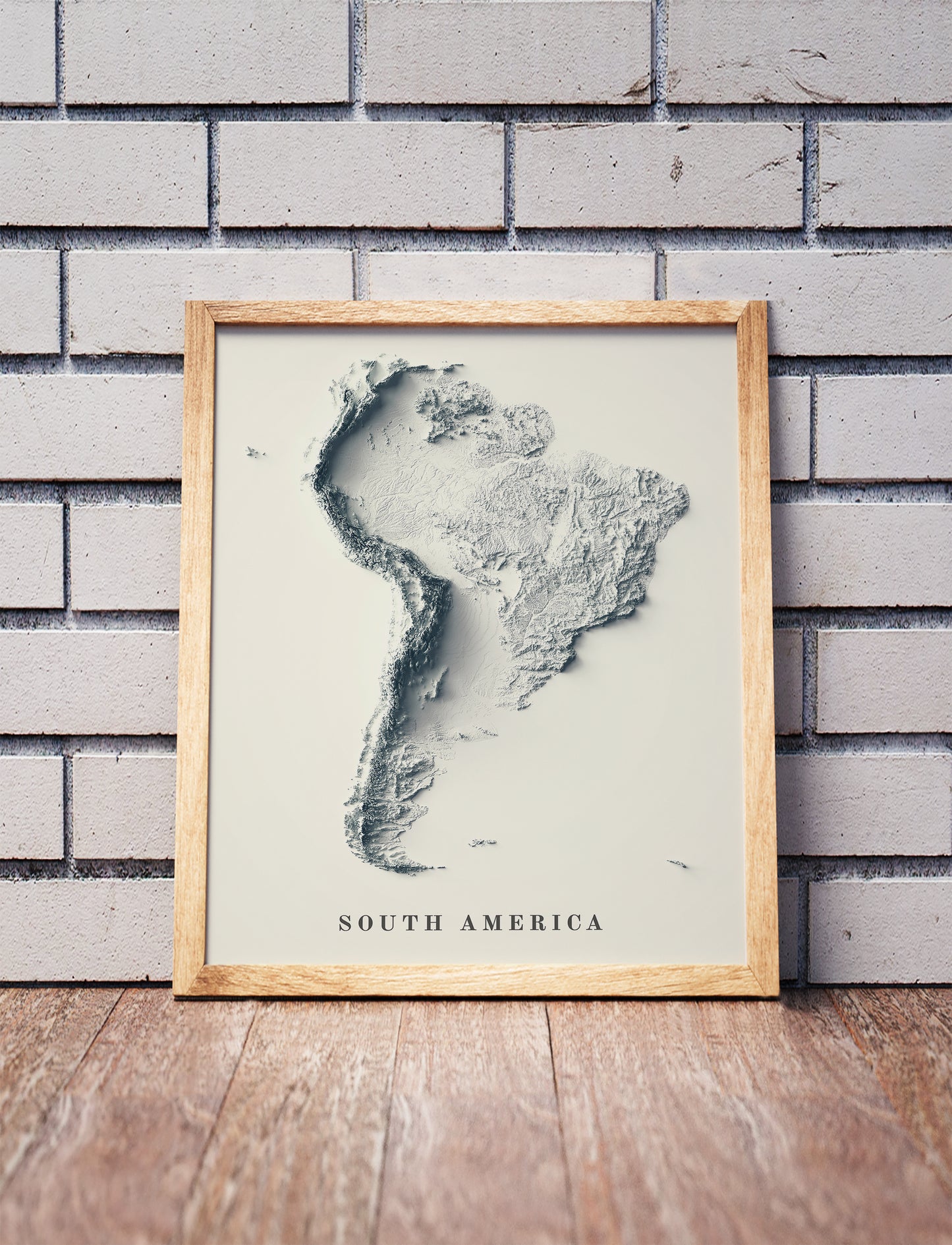 South America shaded relief map in a wooden frame. Realistic terrain art print for home decor, displayed against a brick wall