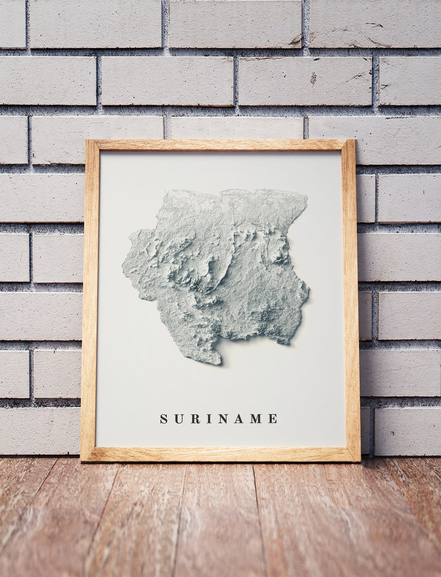 Suriname shaded relief map in a wooden frame. Realistic terrain art print for home decor, displayed against a brick wall