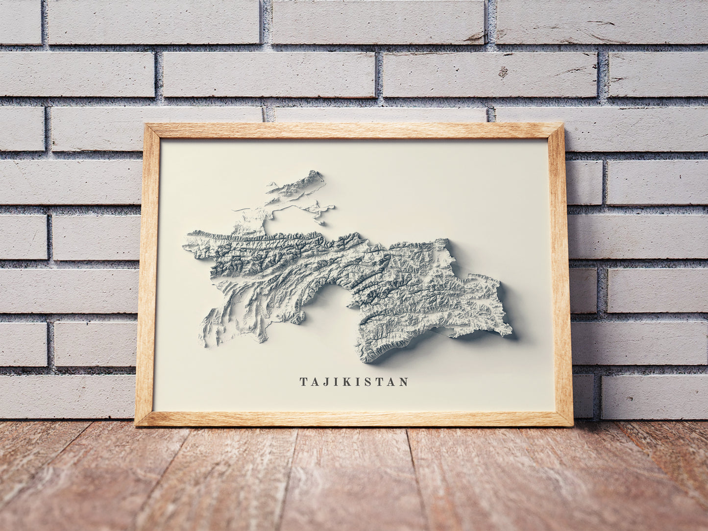 Tajikistan shaded relief map in a wooden frame. Realistic terrain art print for home decor, displayed against a brick wall