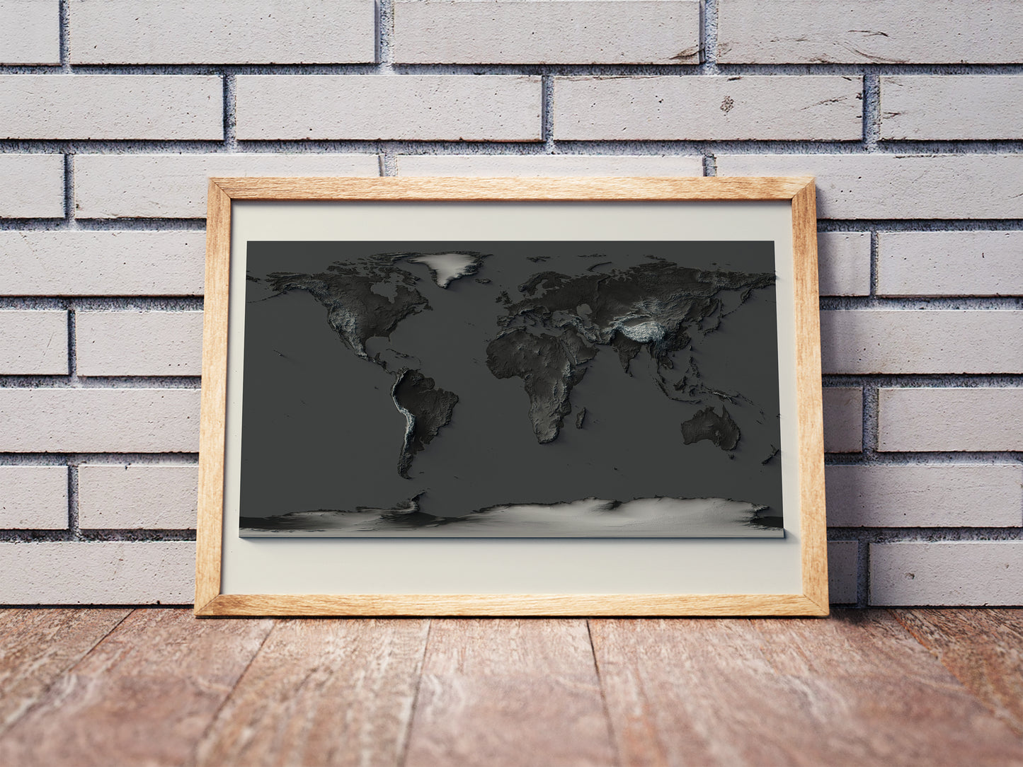 World shaded relief map with dark tone in a wooden frame. Realistic terrain art print for home decor, displayed against a brick wall