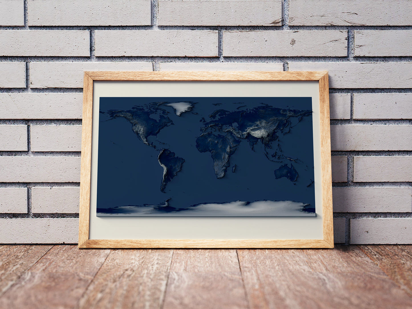 World shaded relief map with dark tones in a wooden frame. Realistic terrain art print for home decor, displayed against a brick wall