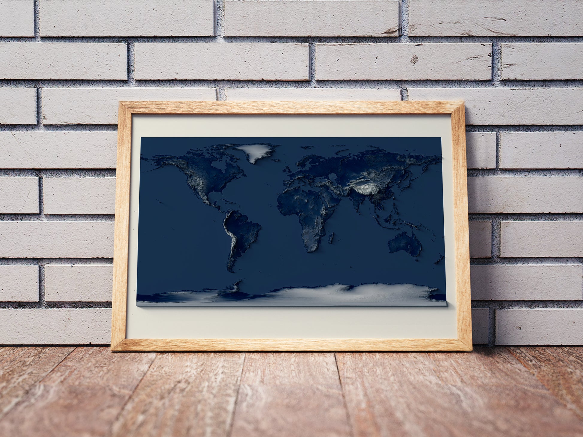 World shaded relief map with dark tones in a wooden frame. Realistic terrain art print for home decor, displayed against a brick wall