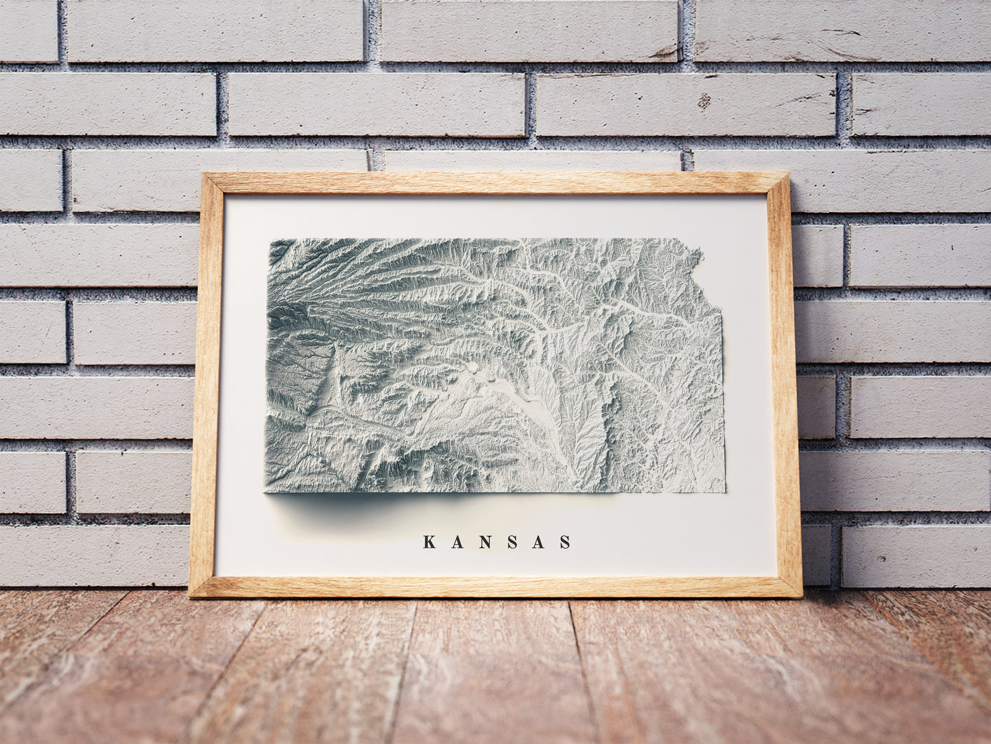 Kansas shaded relief map in a wooden frame. Realistic terrain art print for home decor, displayed against a brick wall
