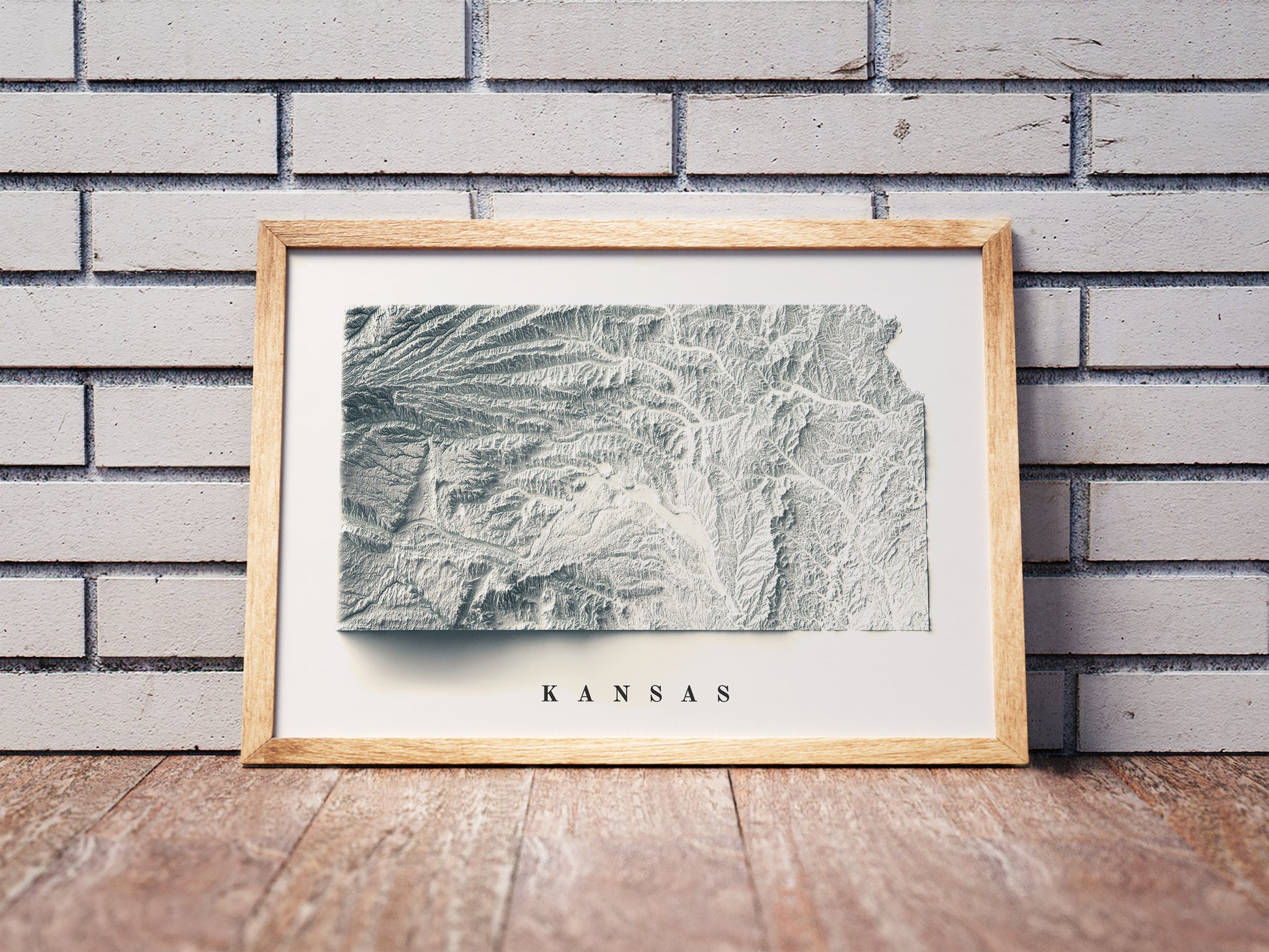 Kansas shaded relief map in a wooden frame. Realistic terrain art print for home decor, displayed against a brick wall