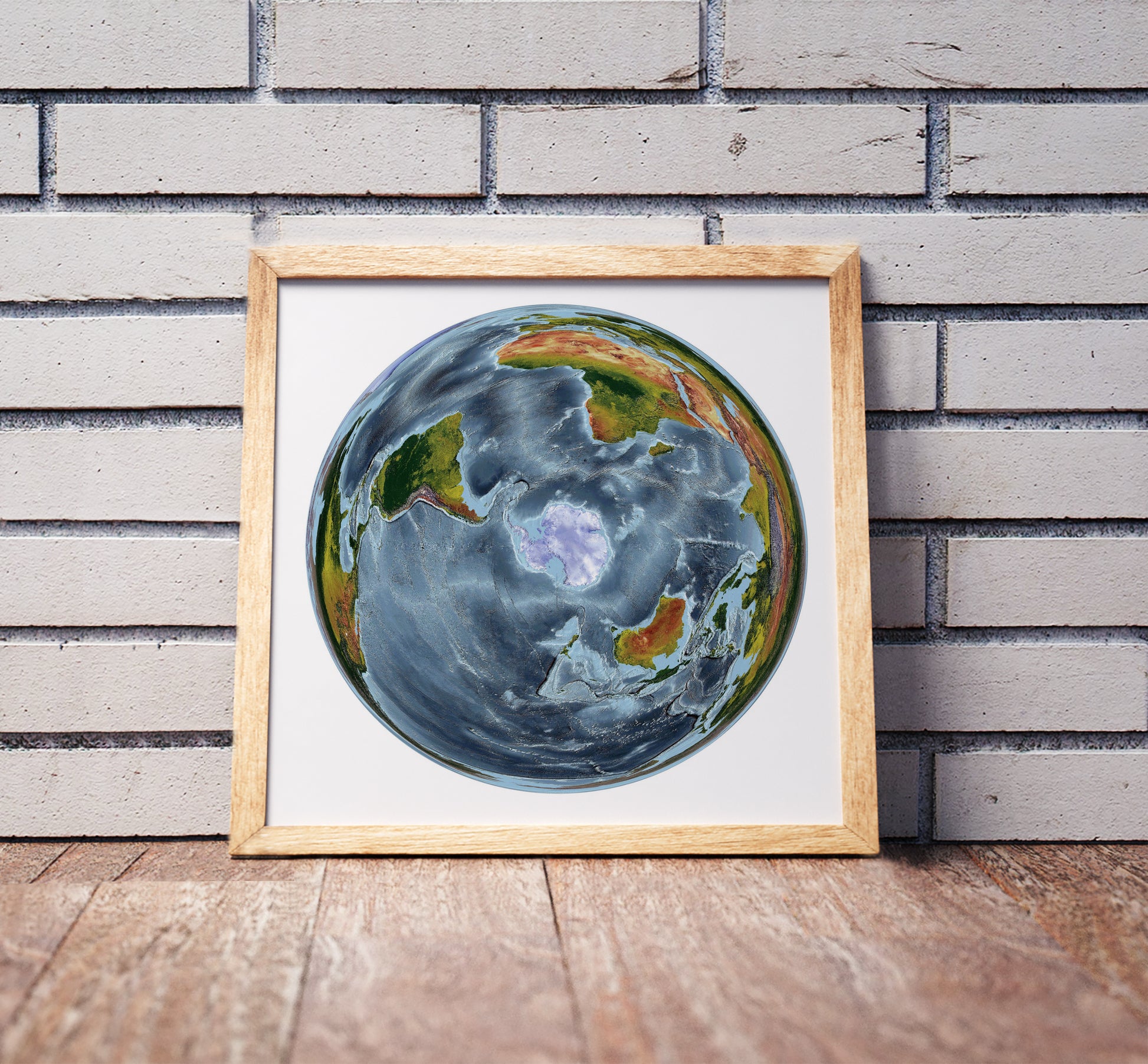 WGS 1984 Grid South projection shaded relief world map in a wooden frame. Realistic terrain art print for home decor, displayed against a brick wall