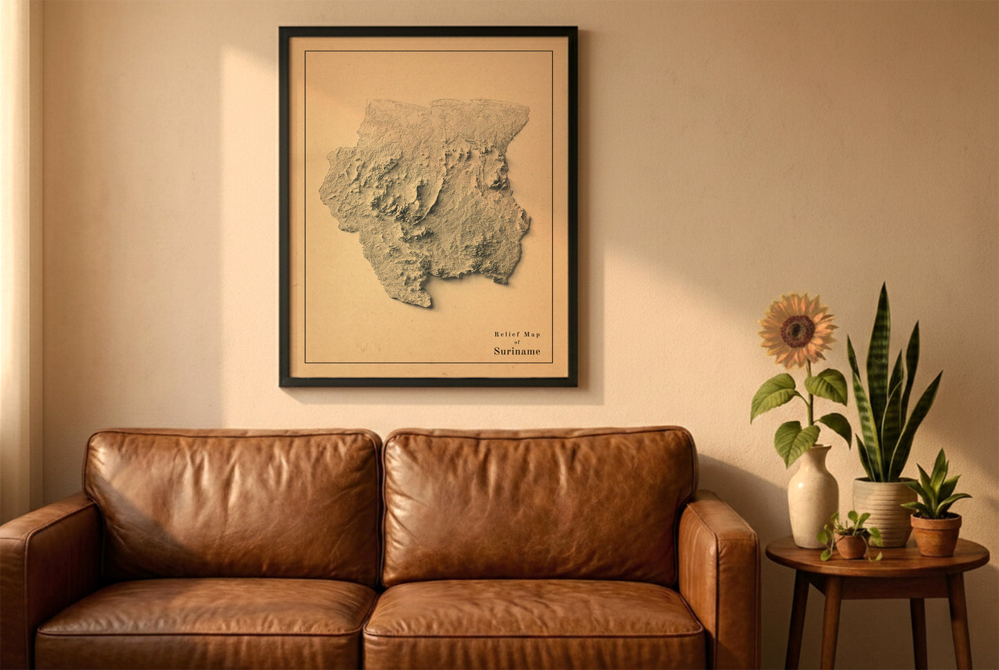 Suriname vintage relief map in a black frame above a brown leather sofa. Boho living room with warm sunlight, sunflower, and indoor plants on a wooden side table