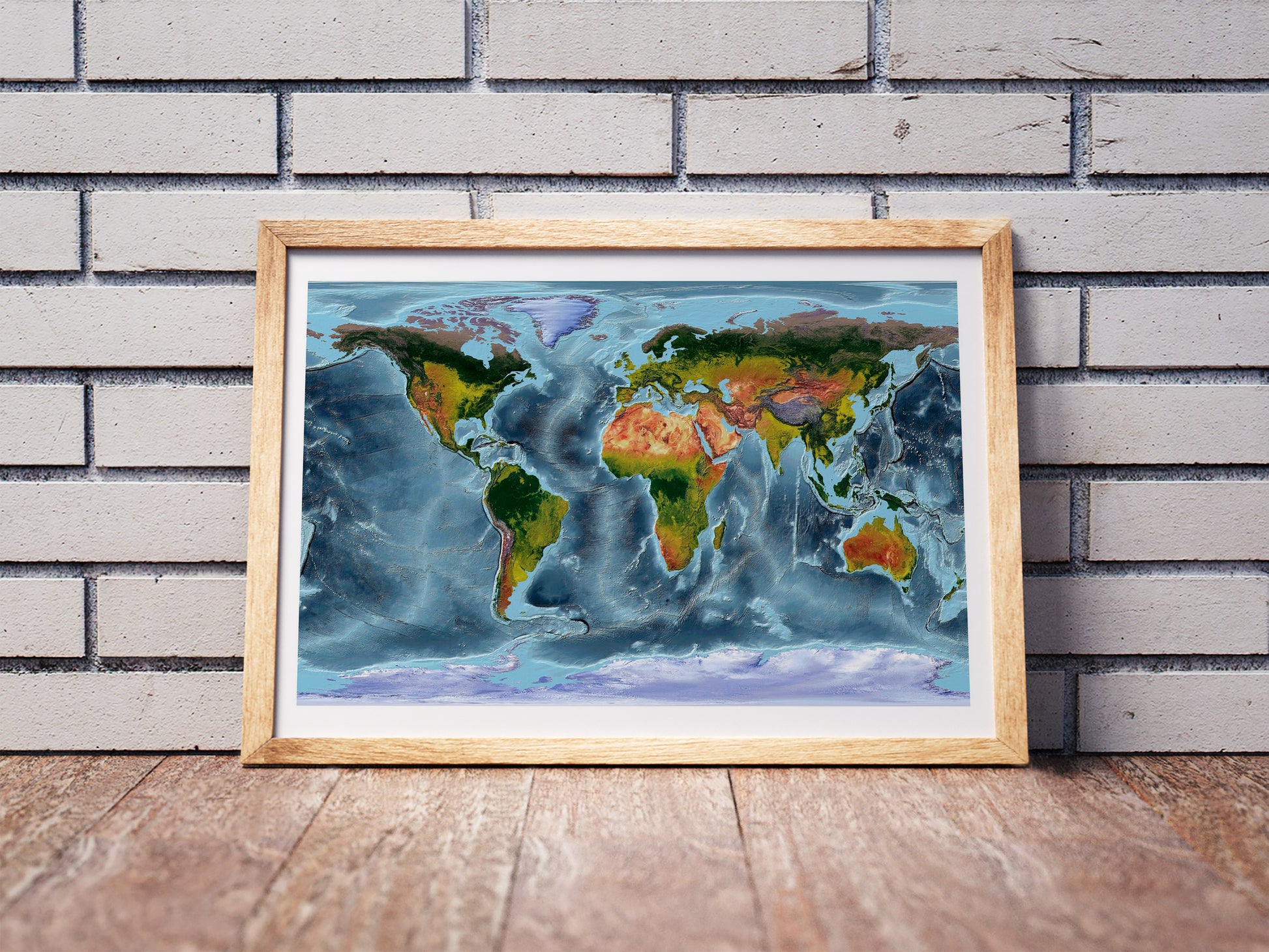 WGS 84 shaded relief world map in a wooden frame. Realistic terrain art print for home decor, displayed against a brick wall
