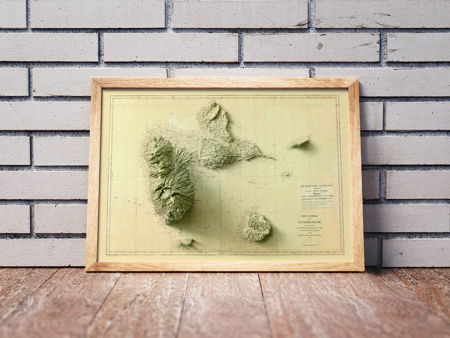 Vintage Guadeloupe, France, shaded relief map in a wooden frame. Realistic terrain art print for home decor, displayed against a brick wall