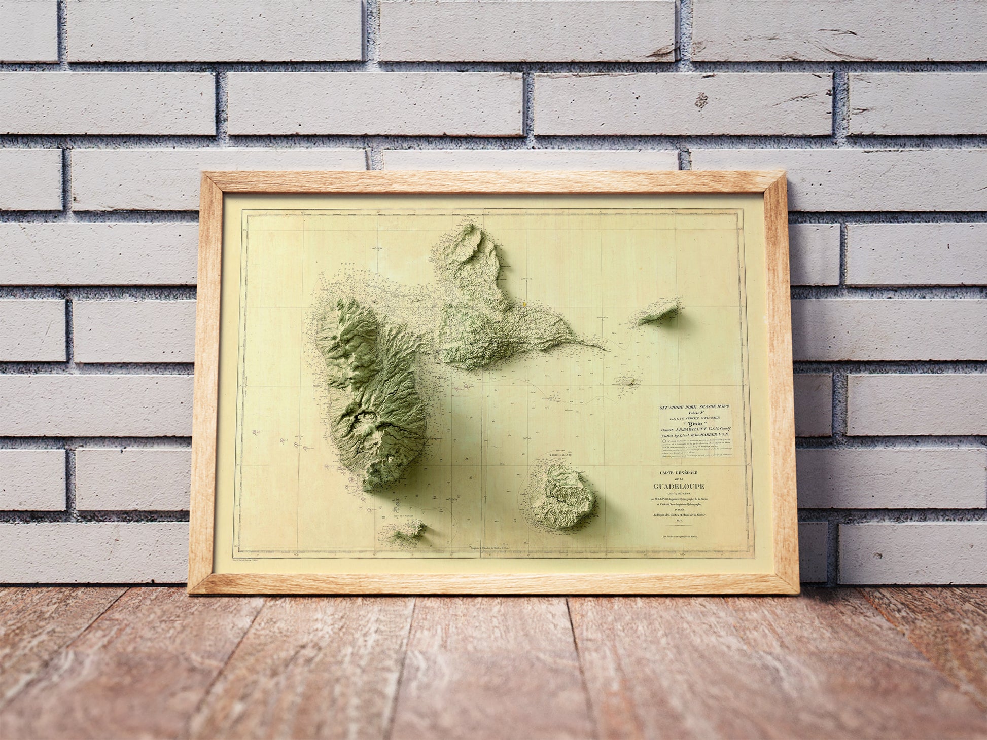 Vintage Guadeloupe, France, shaded relief map in a wooden frame. Realistic terrain art print for home decor, displayed against a brick wall