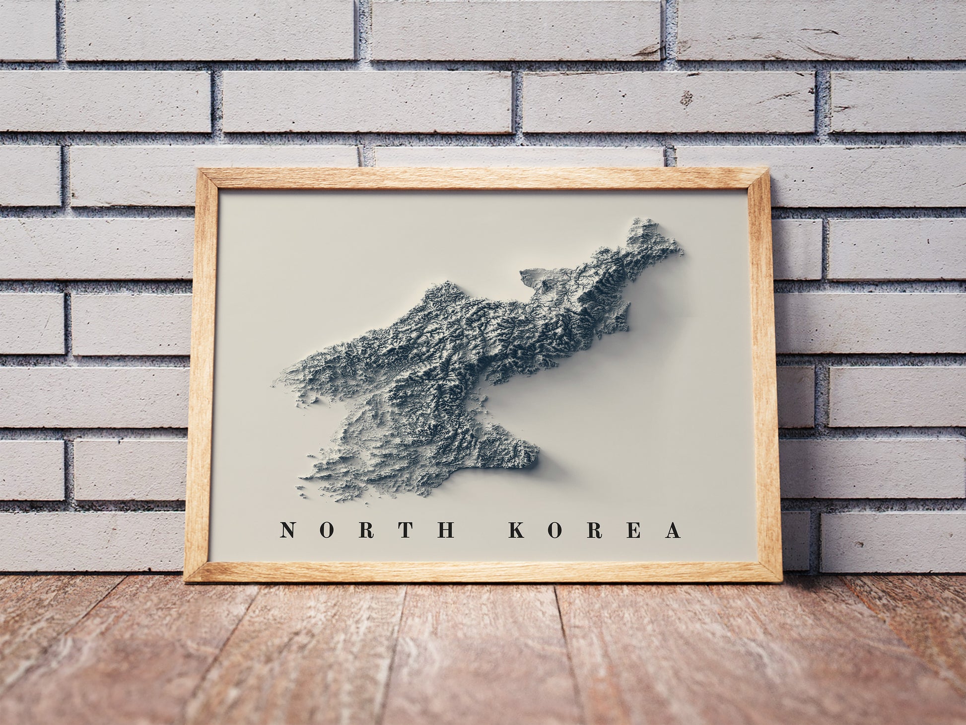 North Korea shaded relief map in a wooden frame. Realistic terrain art print for home decor, displayed against a brick wall