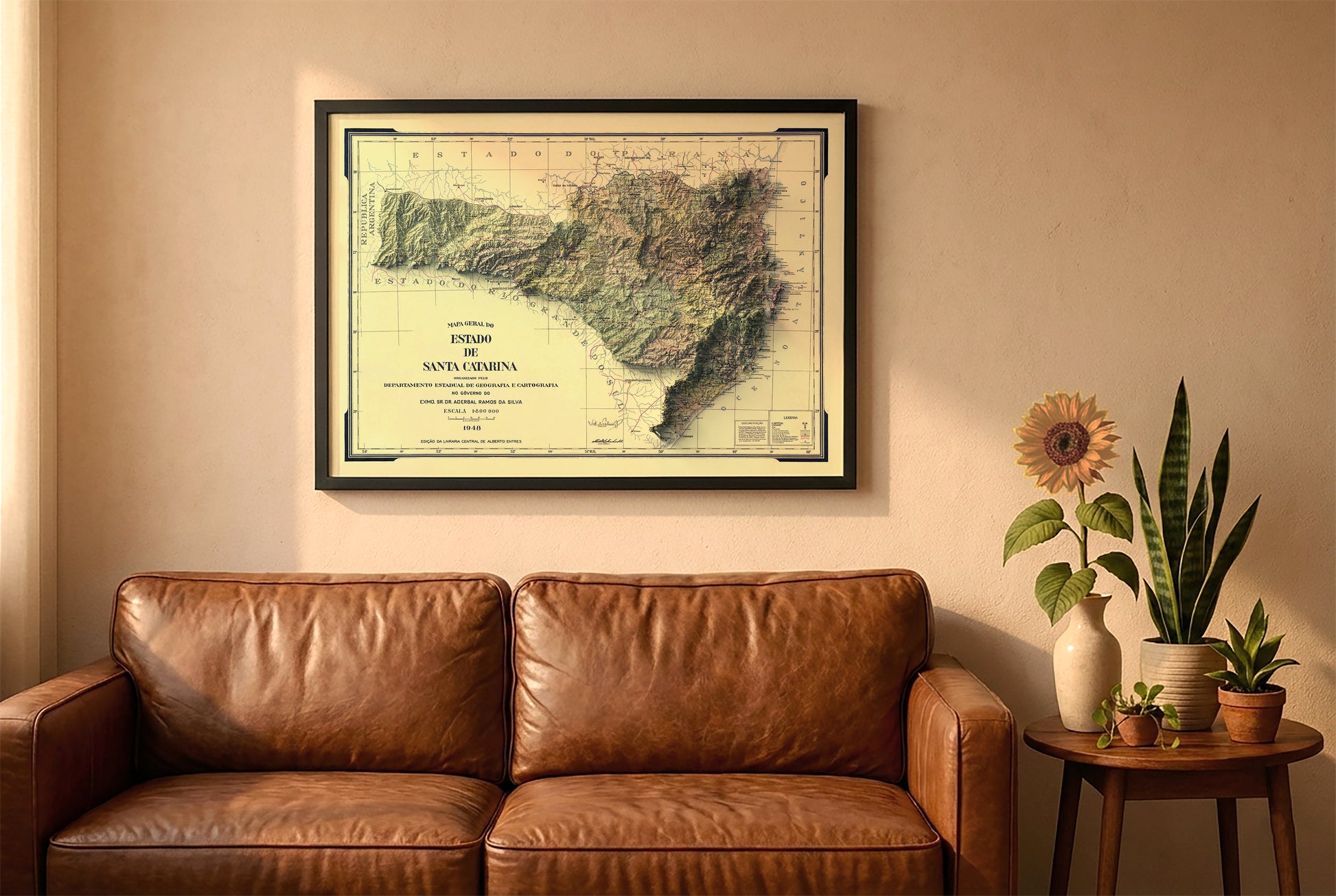 Santa Catarina, Brazil, vintage relief map in a black frame above a brown leather sofa. Boho living room with warm sunlight, sunflower, and indoor plants on a wooden side table