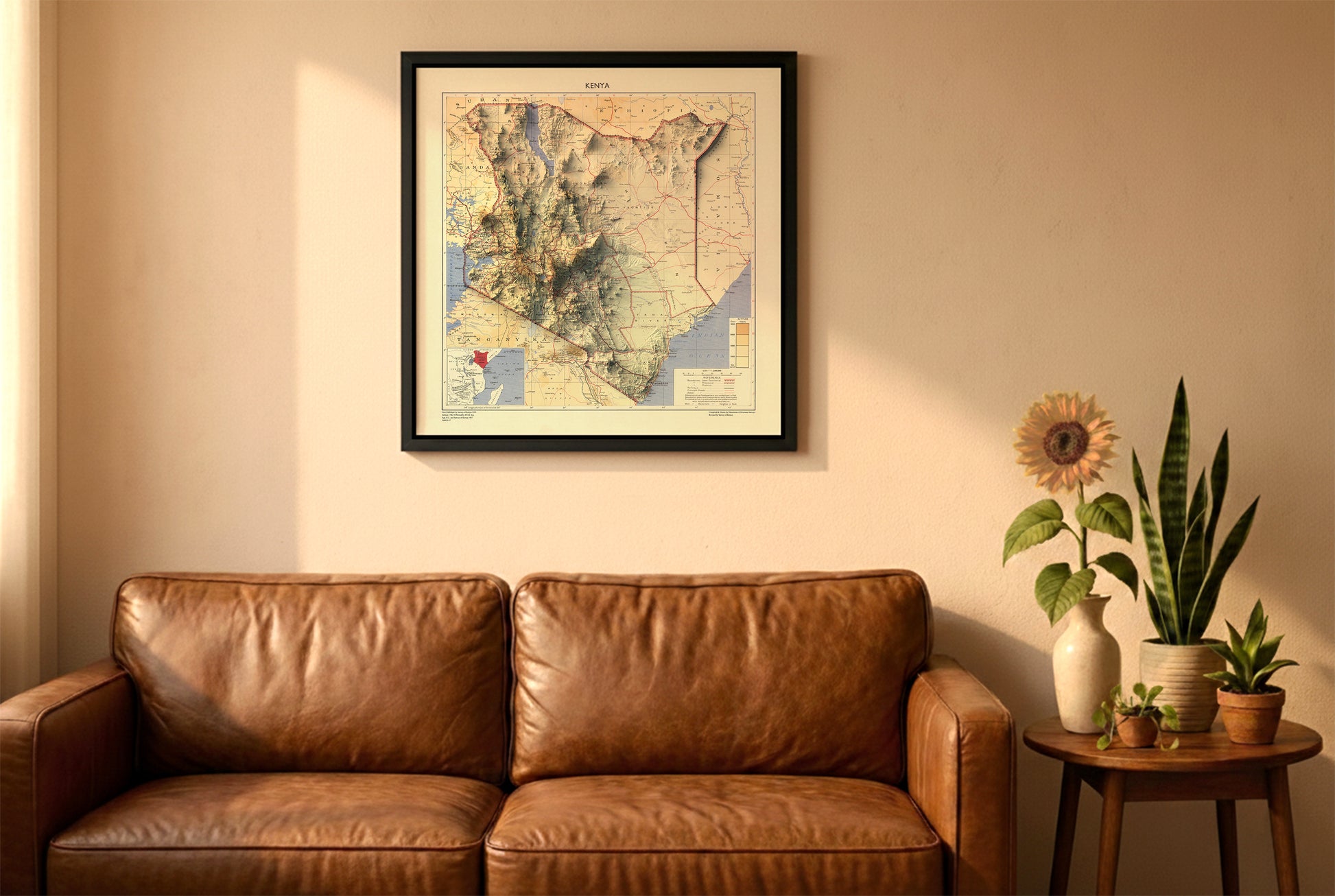 Kenya vintage relief map in a black frame above a brown leather sofa. Boho living room with warm sunlight, sunflower, and indoor plants on a wooden side table