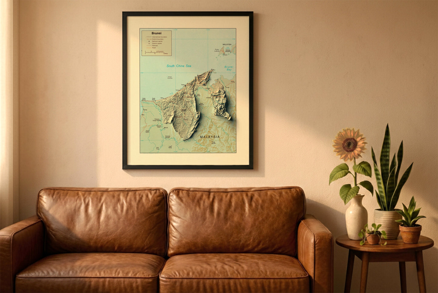 Brunei vintage relief map in a black frame above a brown leather sofa. Boho living room with warm sunlight, sunflower, and indoor plants on a wooden side table