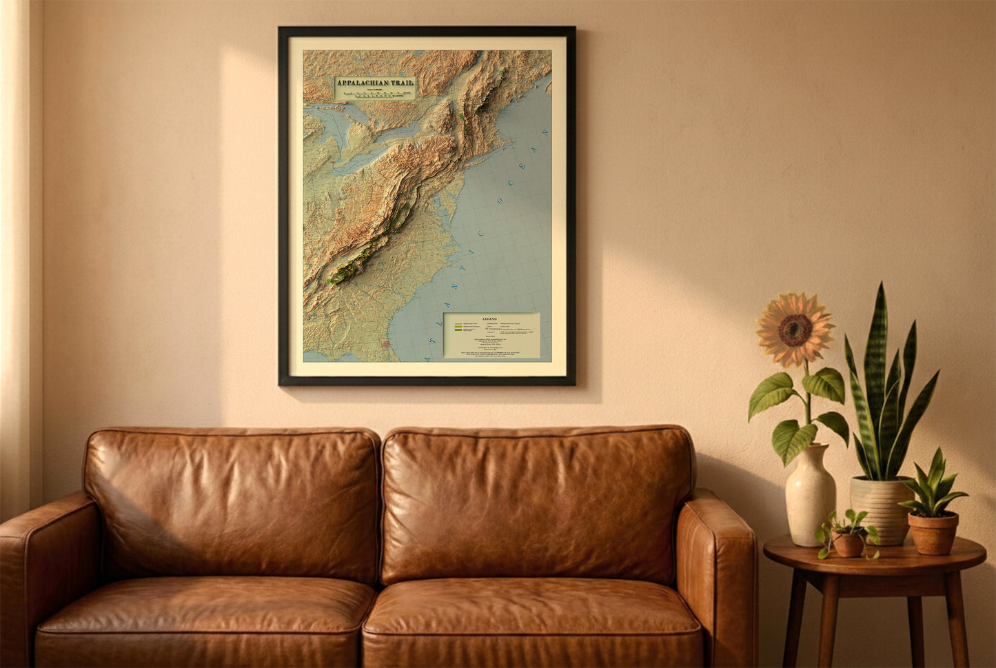 Appalachian Trail vintage relief map in a black frame above a brown leather sofa. Boho living room with warm sunlight, sunflower, and indoor plants on a wooden side table