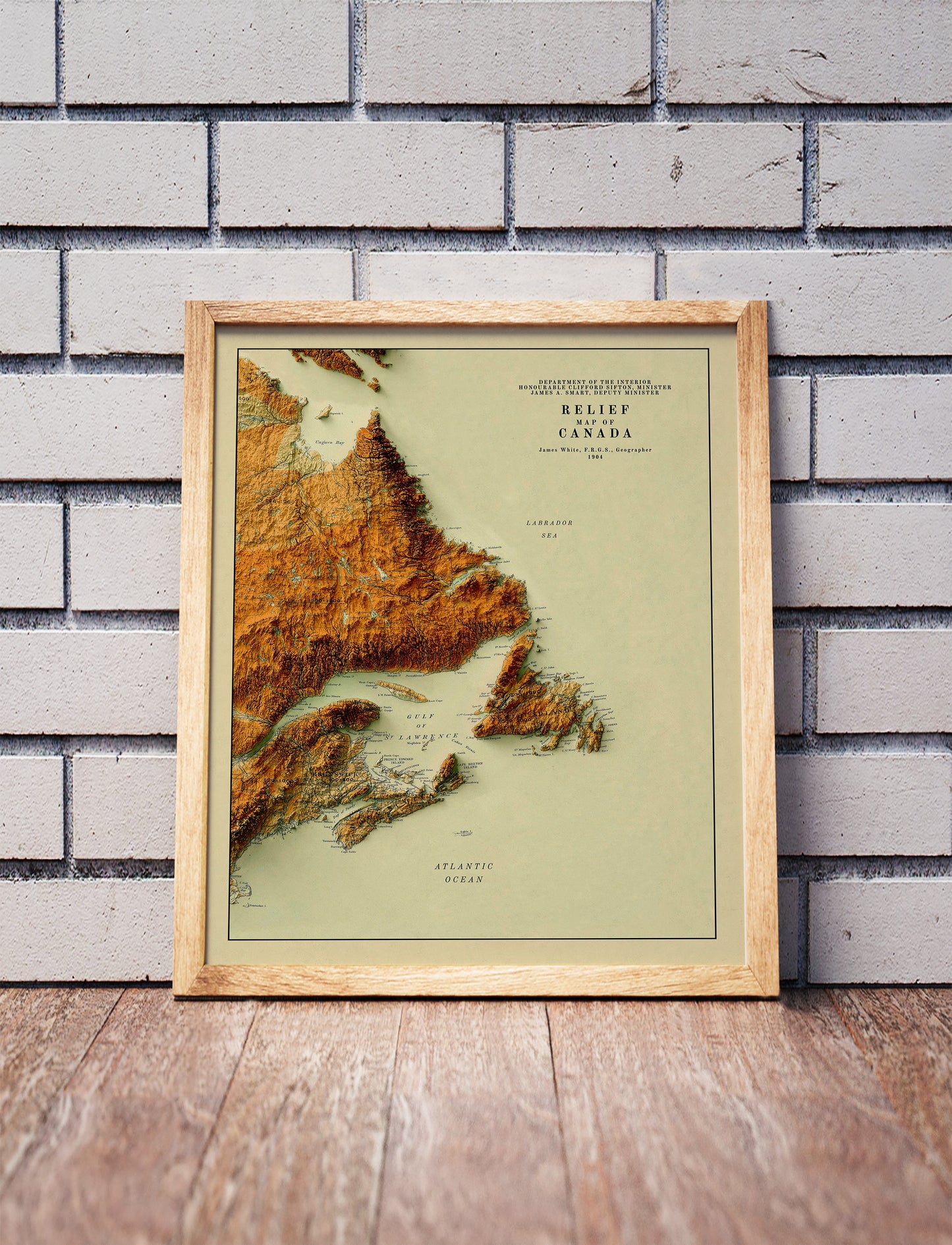 Vintage Atlantic Canada shaded relief map in a wooden frame. Realistic terrain art print for home decor, displayed against a brick wall
