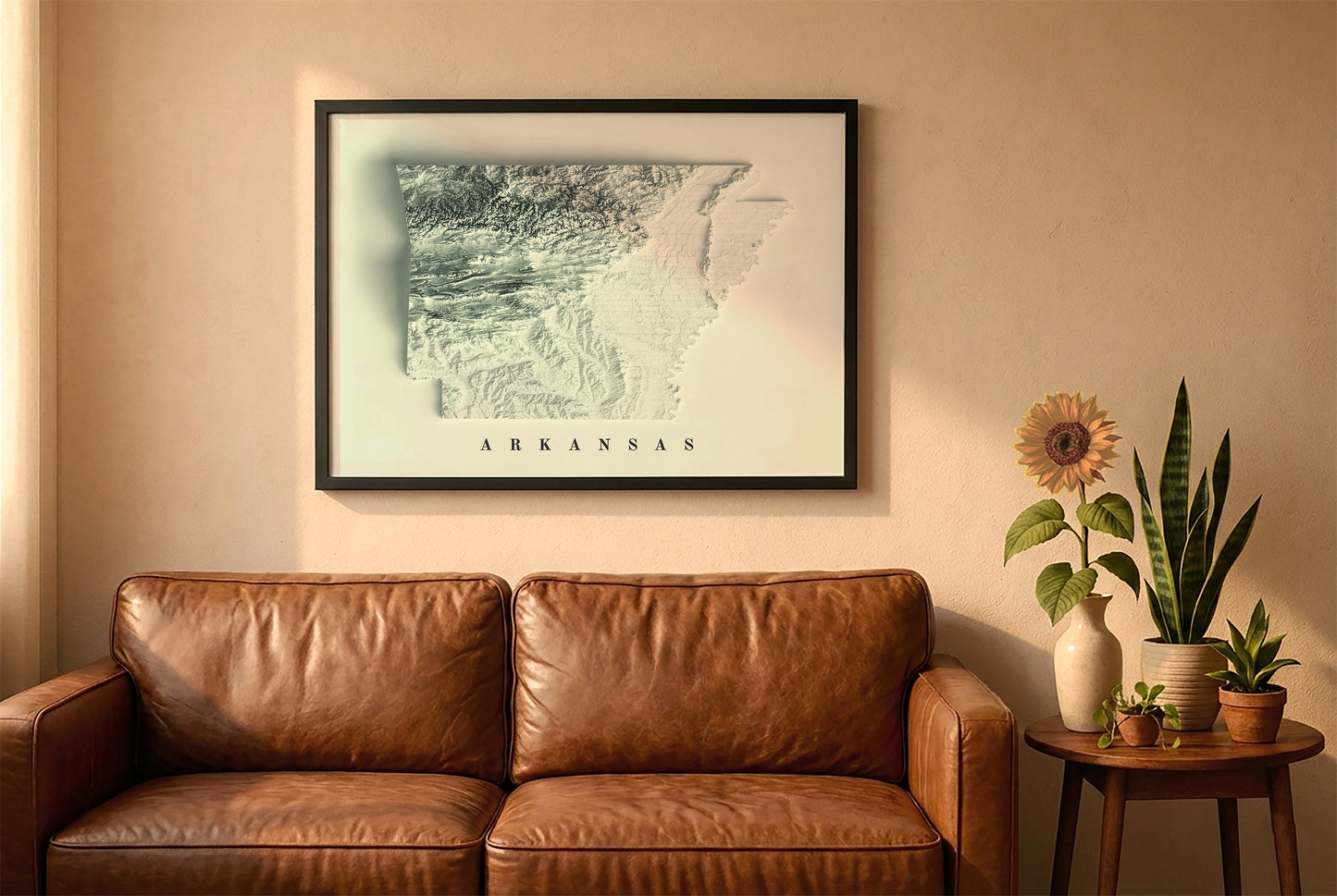 Arkansas relief map in a black frame above a brown leather sofa. Boho living room with warm sunlight, sunflower, and indoor plants on a wooden side table