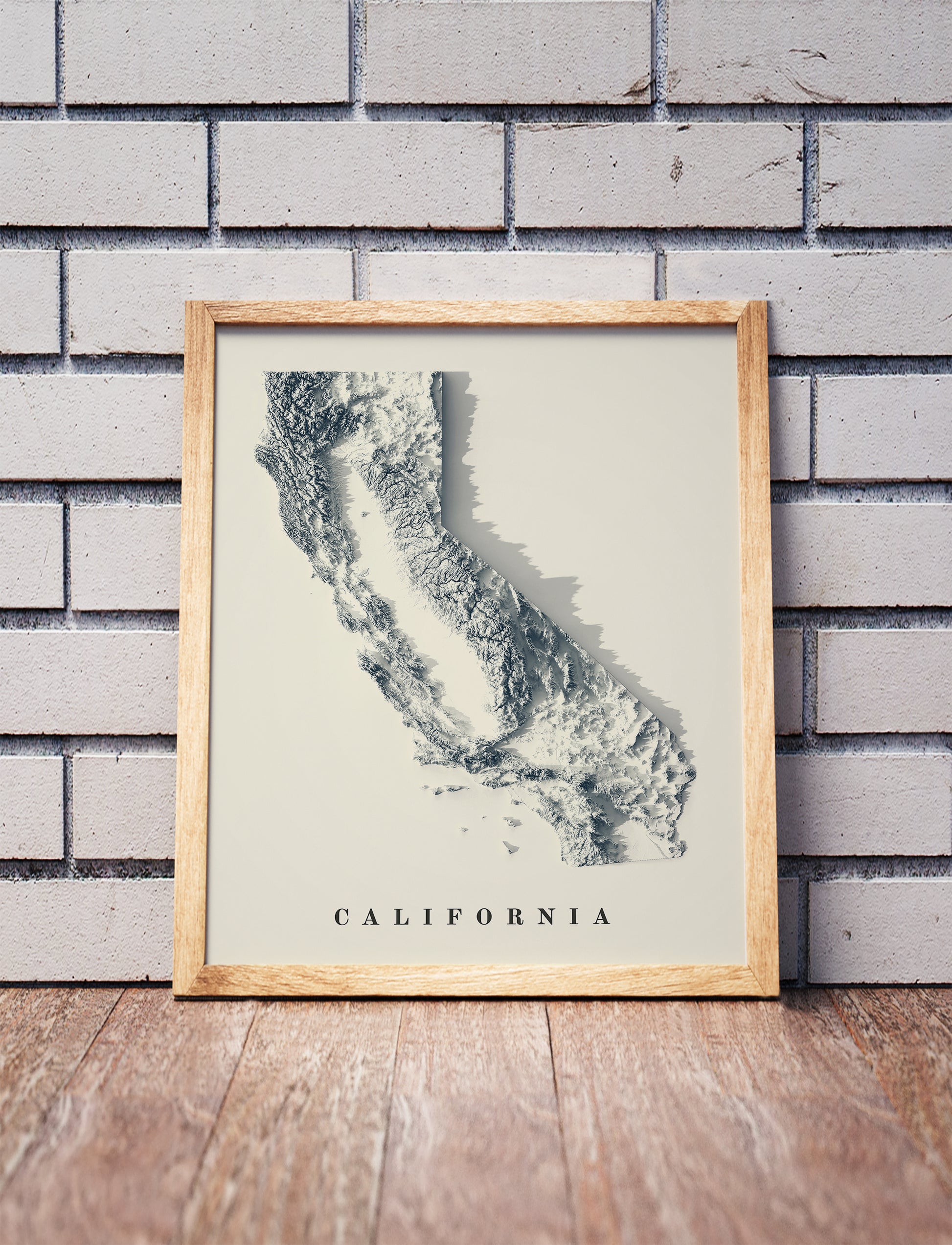 California shaded relief map in a wooden frame. Realistic terrain art print for home decor, displayed against a brick wall