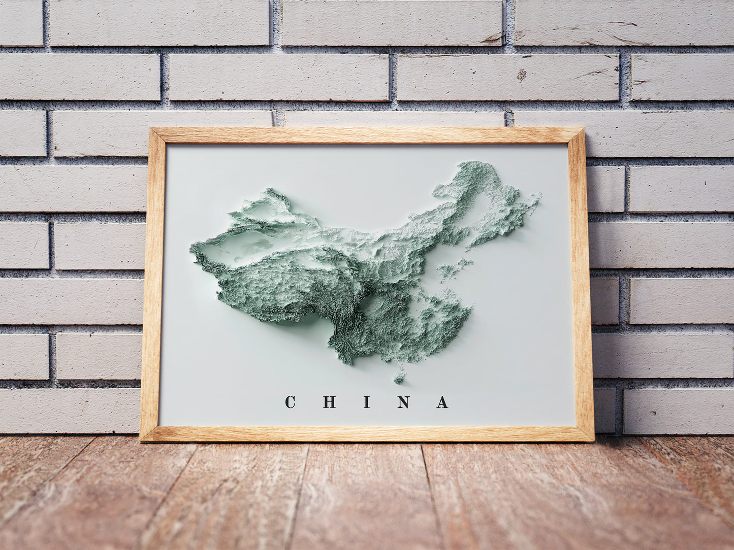 China shaded relief map in a wooden frame. Realistic terrain art print for home decor, displayed against a brick wall
