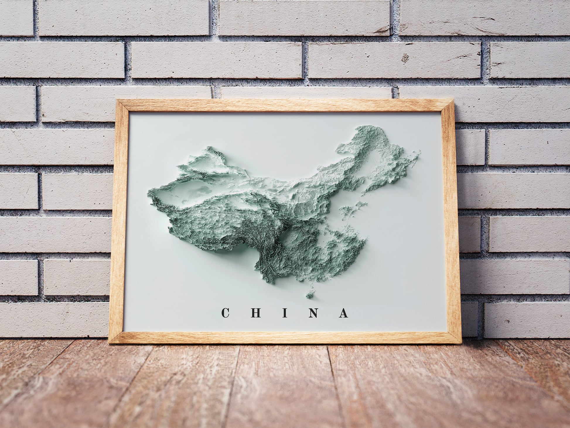 China shaded relief map in a wooden frame. Realistic terrain art print for home decor, displayed against a brick wall