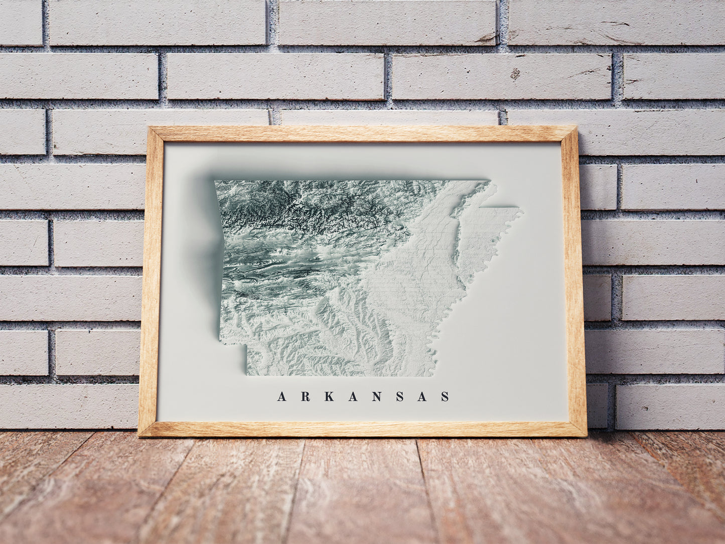 Arkansas shaded relief map in a wooden frame. Realistic terrain art print for home decor, displayed against a brick wall