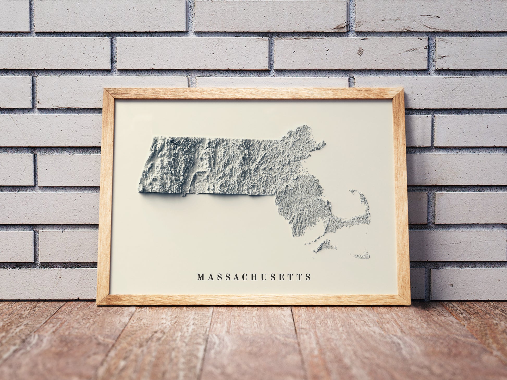 Massachusetts shaded relief map in a wooden frame. Realistic terrain art print for home decor, displayed against a brick wall