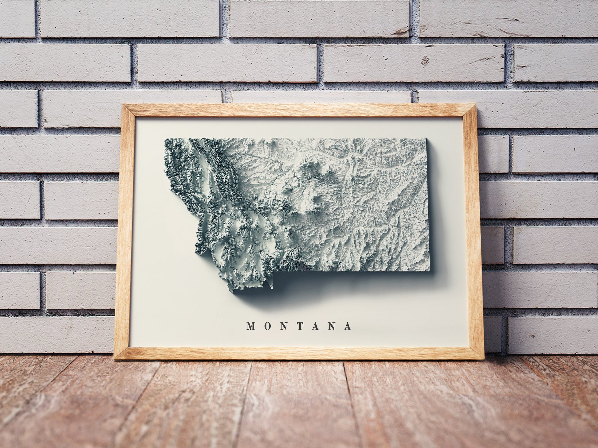 Montana shaded relief map in a wooden frame. Realistic terrain art print for home decor, displayed against a brick wall
