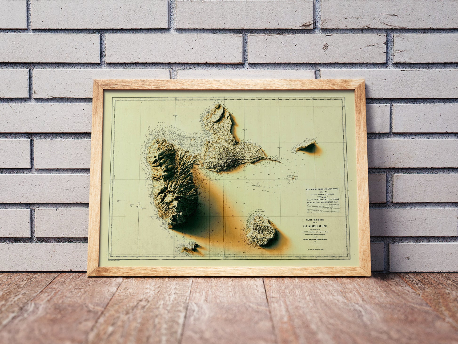 Vintage Guadeloupe, France, shaded relief map in a wooden frame. Realistic terrain art print for home decor, displayed against a brick wall