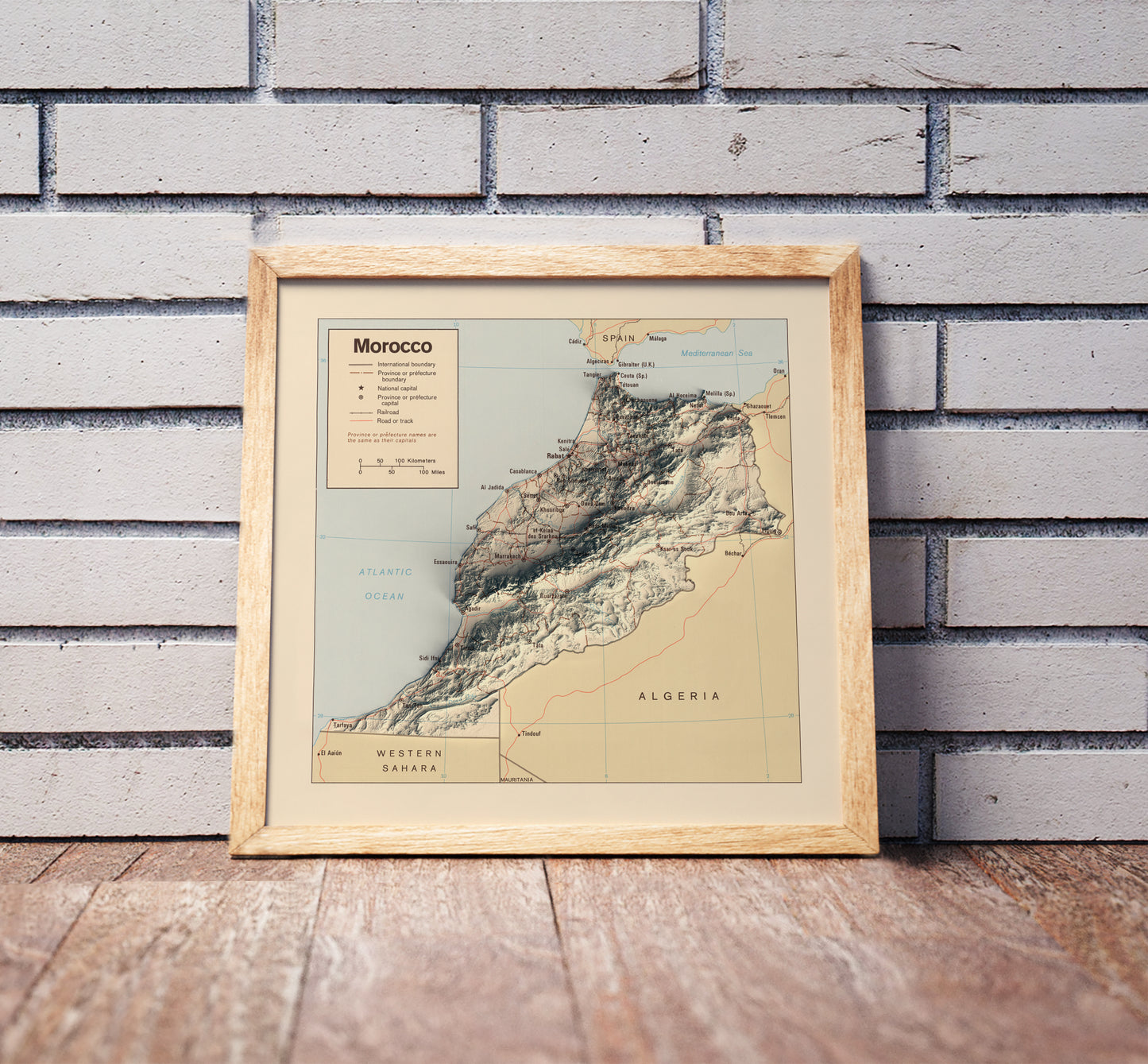 Vintage Morocco shaded relief map in a wooden frame. Realistic terrain art print for home decor, displayed against a brick wall