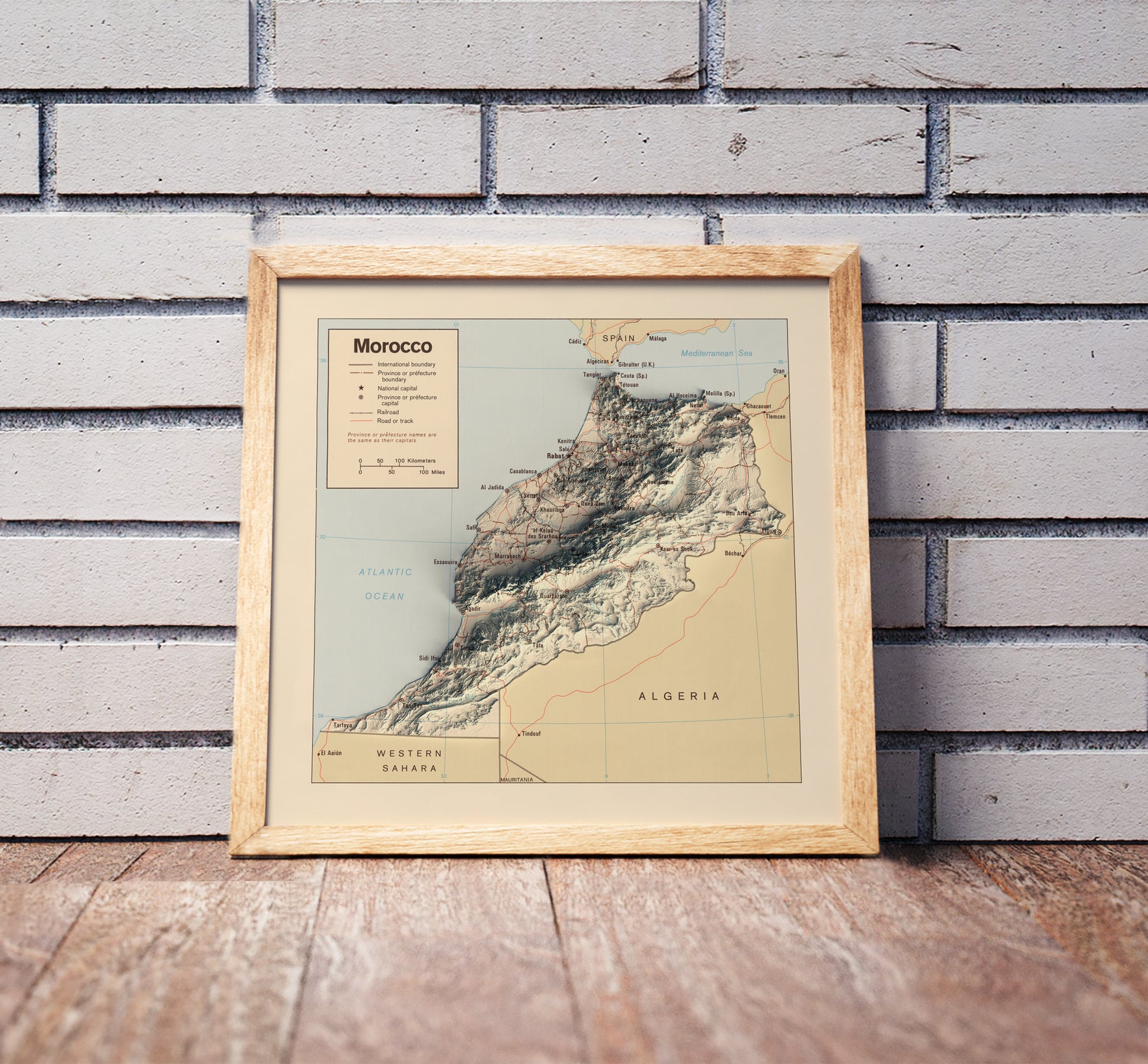 Vintage Morocco shaded relief map in a wooden frame. Realistic terrain art print for home decor, displayed against a brick wall