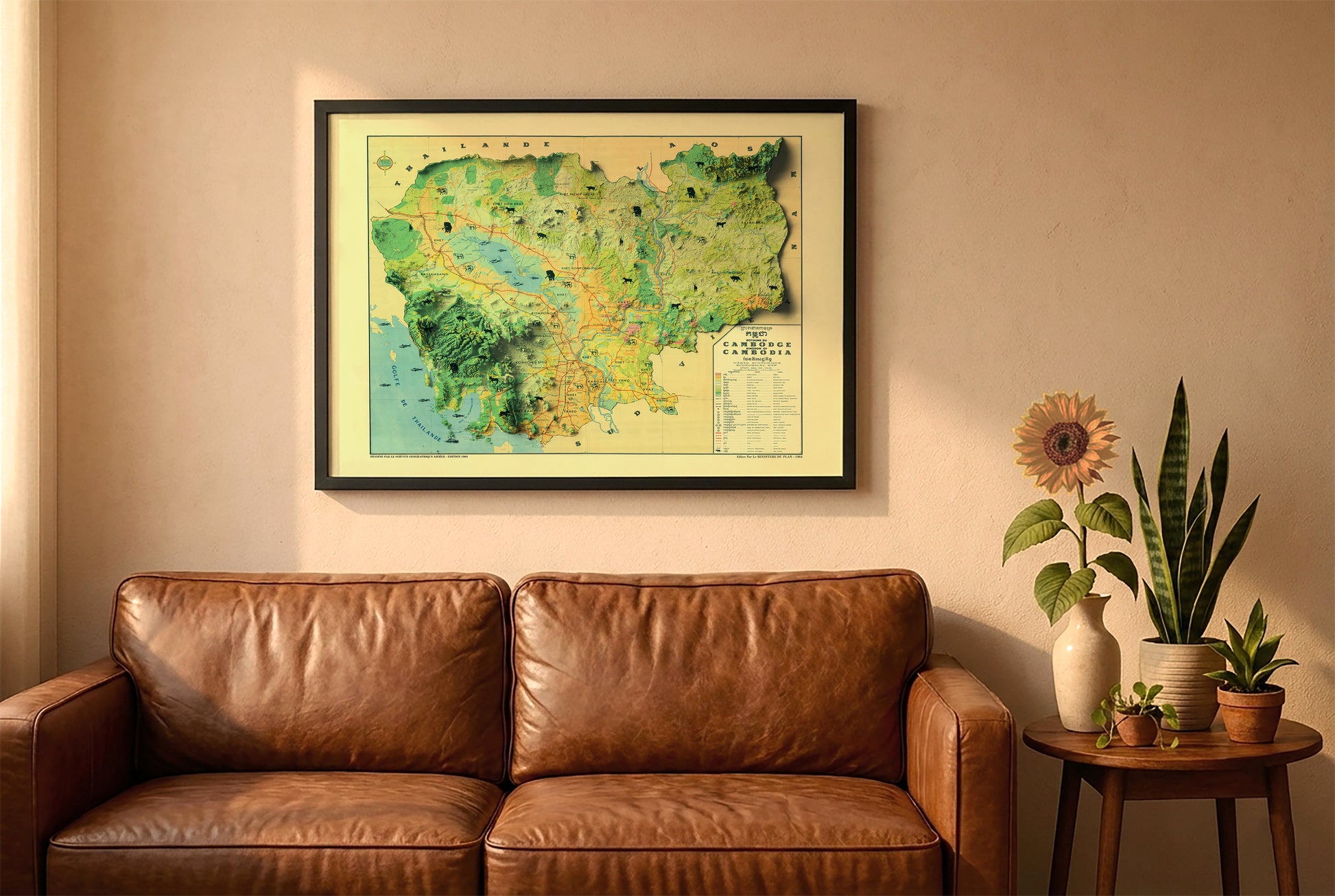 Cambodia vintage relief map in a black frame above a brown leather sofa. Boho living room with warm sunlight, sunflower, and indoor plants on a wooden side table