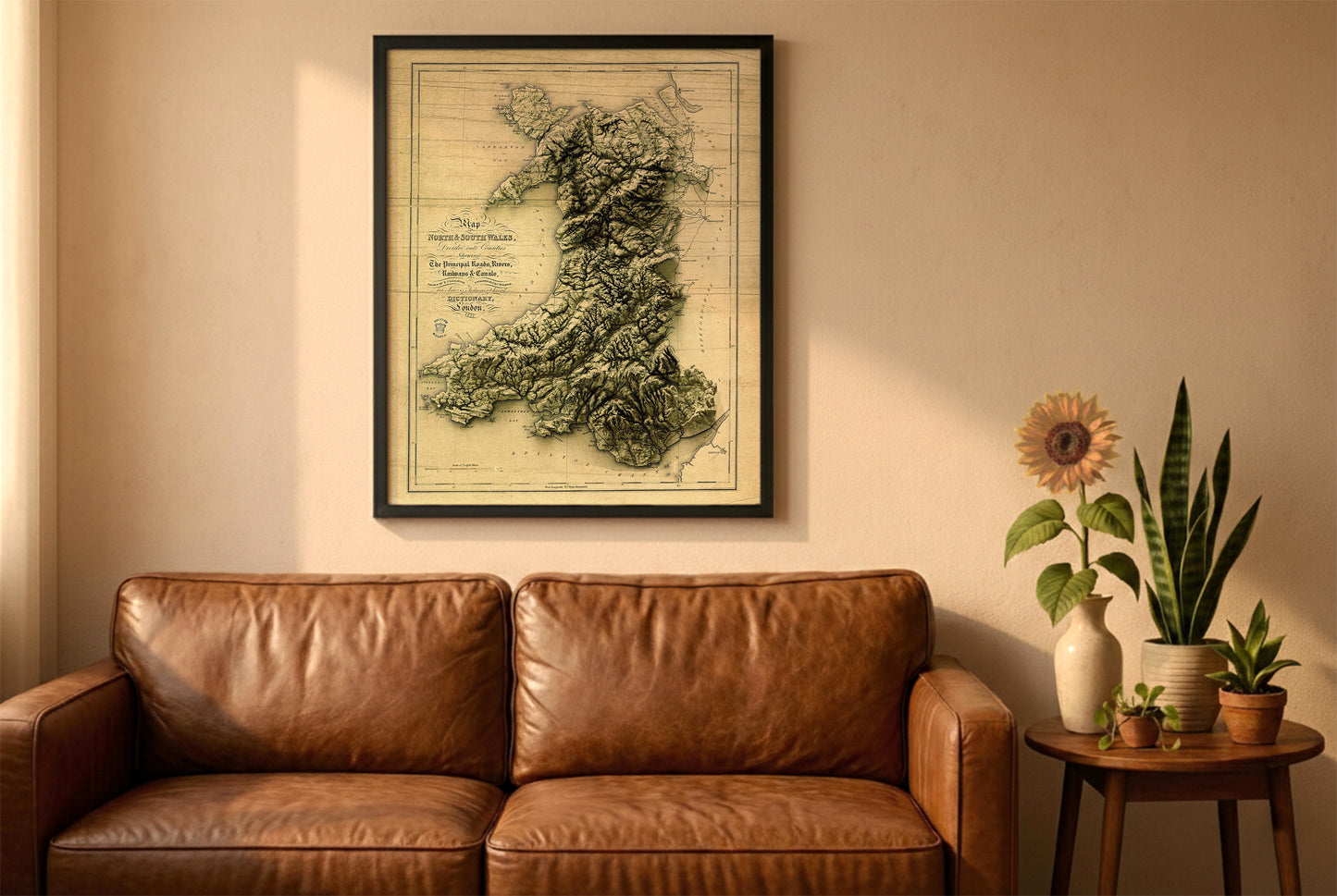 Wales vintage relief map in a black frame above a brown leather sofa. Boho living room with warm sunlight, sunflower, and indoor plants on a wooden side table