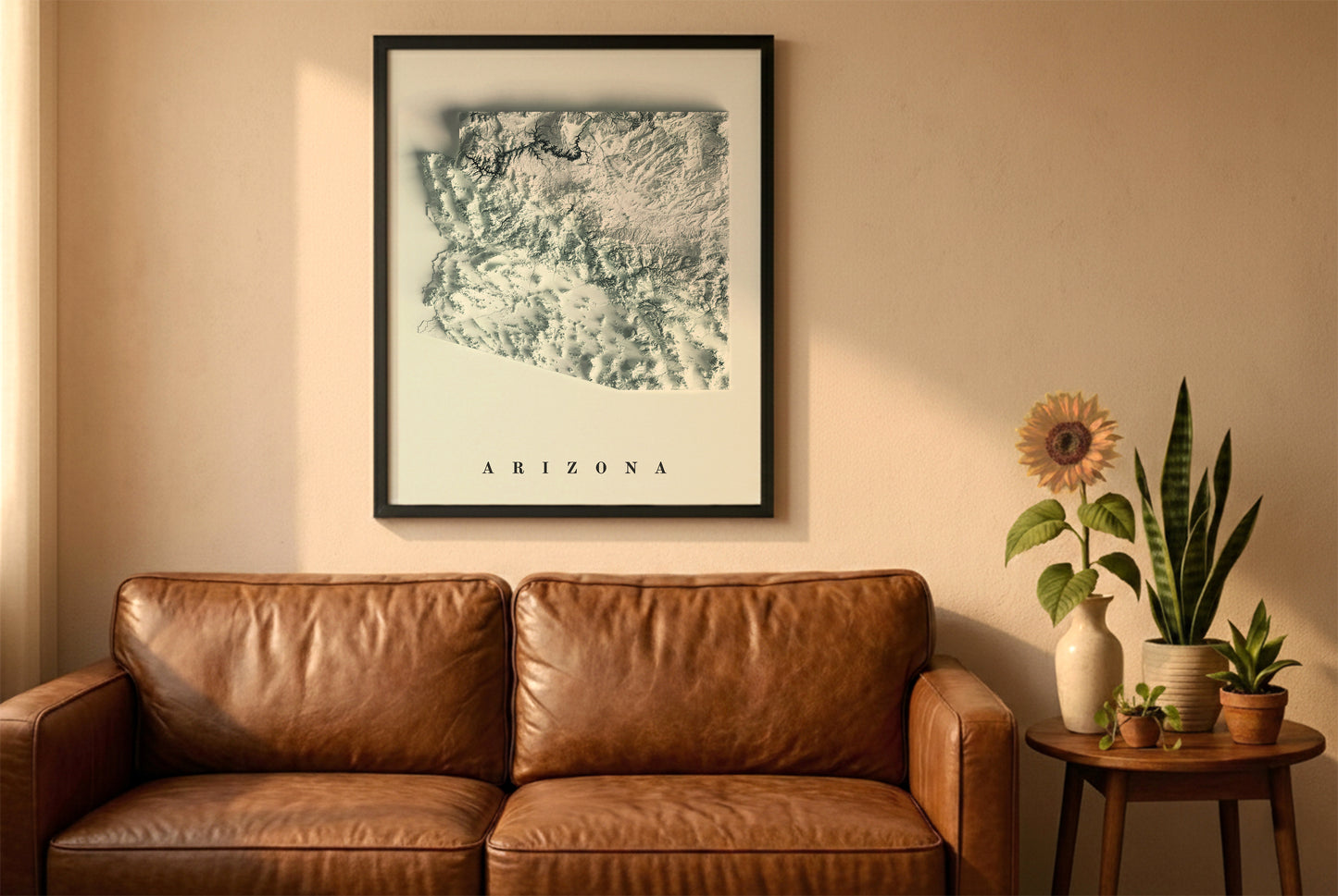 Arizona relief map in a black frame above a brown leather sofa. Boho living room with warm sunlight, sunflower, and indoor plants on a wooden side table