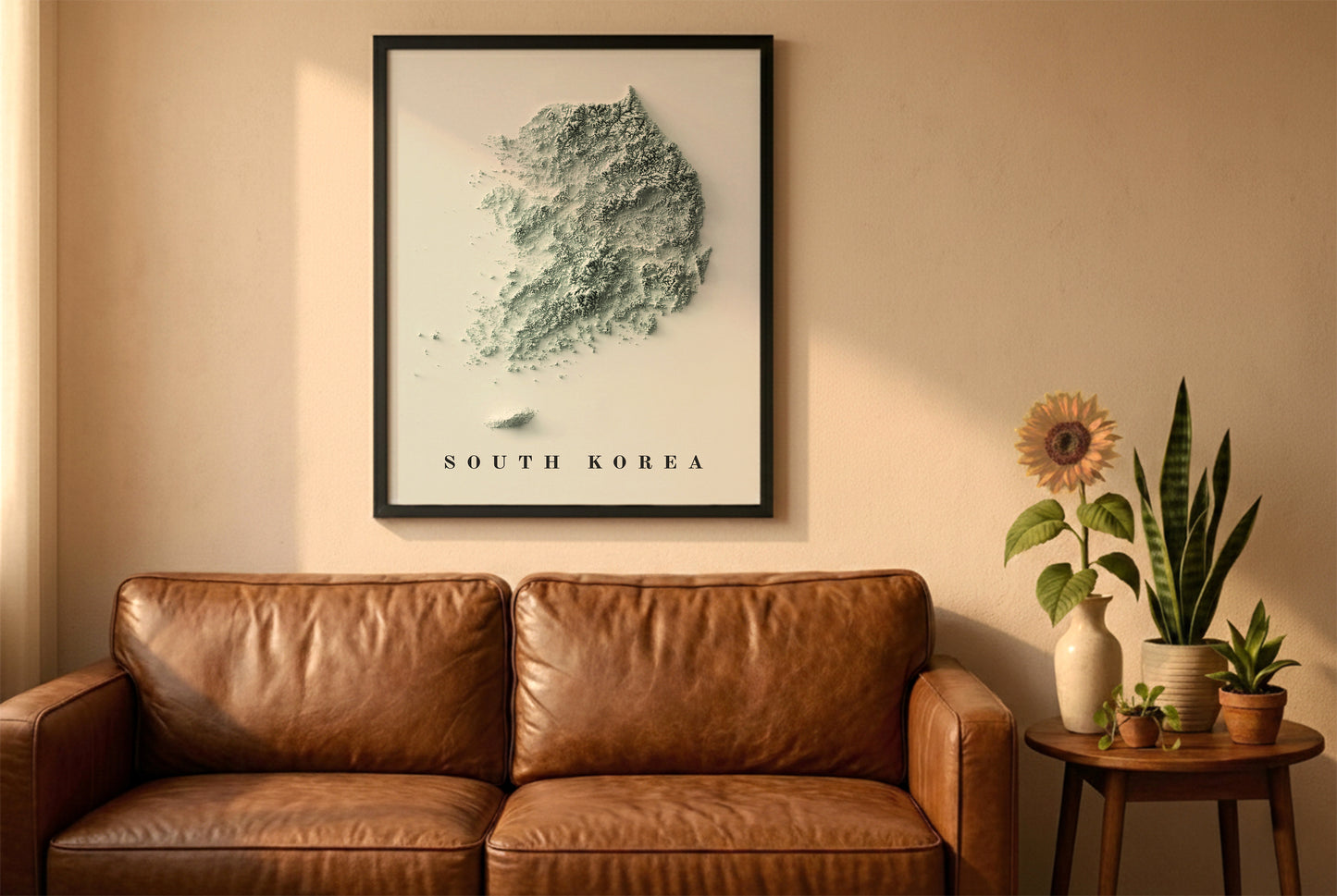 South Korea relief map in a black frame above a brown leather sofa. Boho living room with warm sunlight, sunflower, and indoor plants on a wooden side table