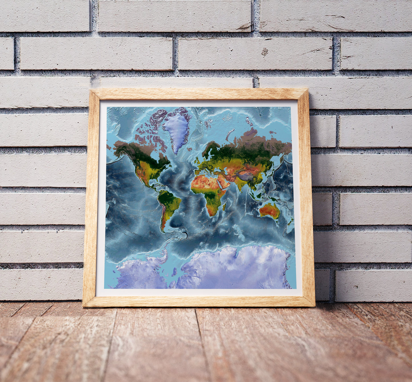 Mercator projection shaded relief world map in a wooden frame. Realistic terrain art print for home decor, displayed against a brick wall