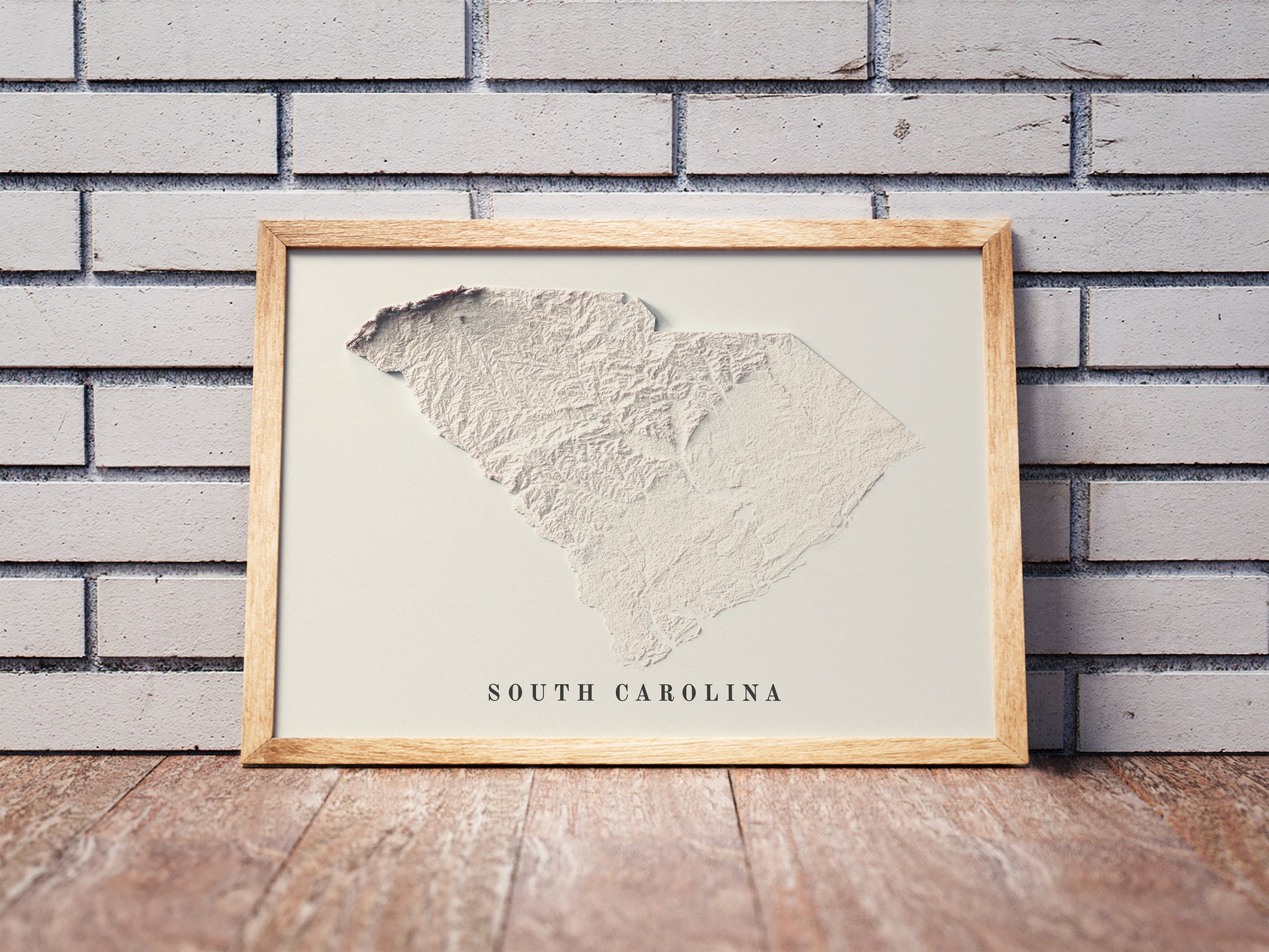 South Carolina shaded relief map in a wooden frame. Realistic terrain art print for home decor, displayed against a brick wall