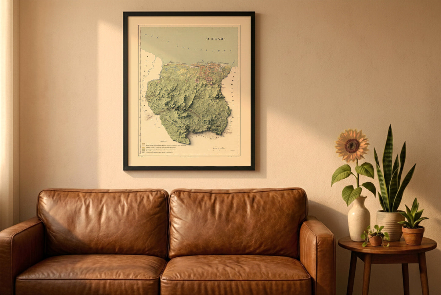 Suriname vintage relief map in a black frame above a brown leather sofa. Boho living room with warm sunlight, sunflower, and indoor plants on a wooden side table