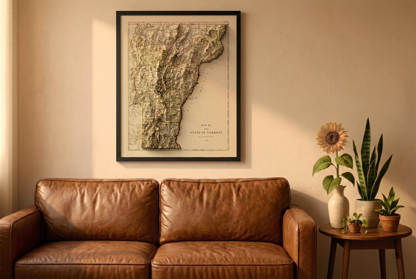 Vermont vintage relief map in a black frame above a brown leather sofa. Boho living room with warm sunlight, sunflower, and indoor plants on a wooden side table