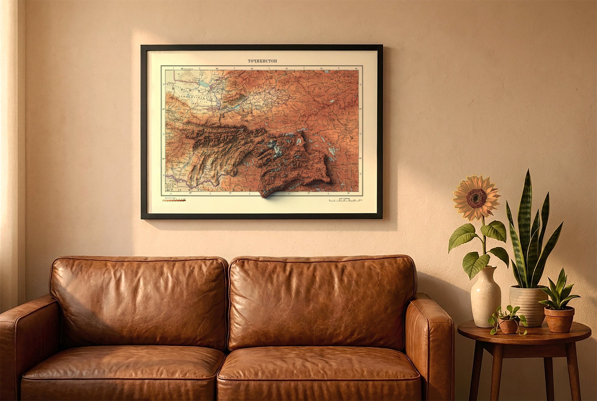 Tajikistan vintage relief map in a black frame above a brown leather sofa. Boho living room with warm sunlight, sunflower, and indoor plants on a wooden side table