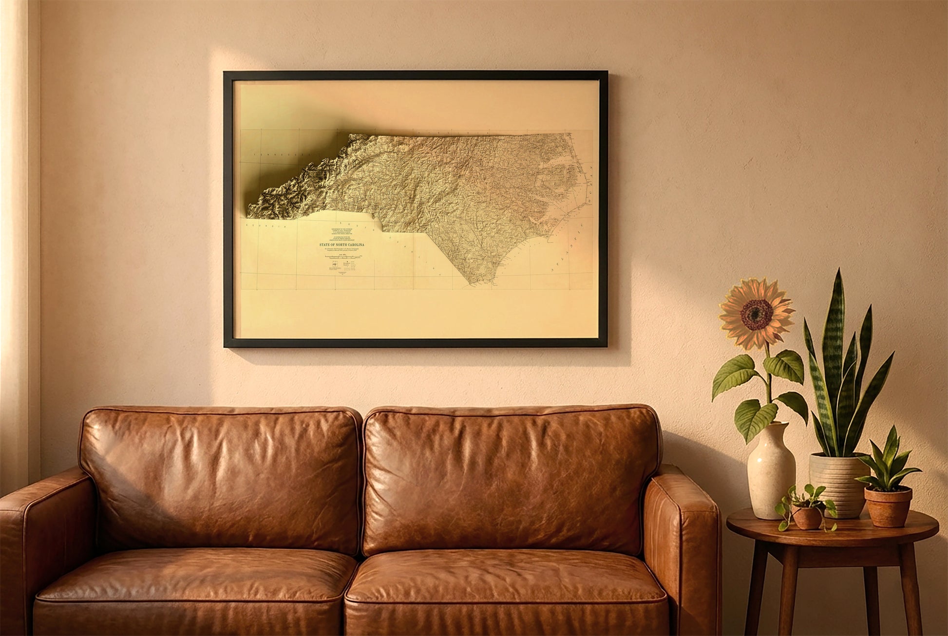 North Carolina vintage relief map in a black frame above a brown leather sofa. Boho living room with warm sunlight, sunflower, and indoor plants on a wooden side table