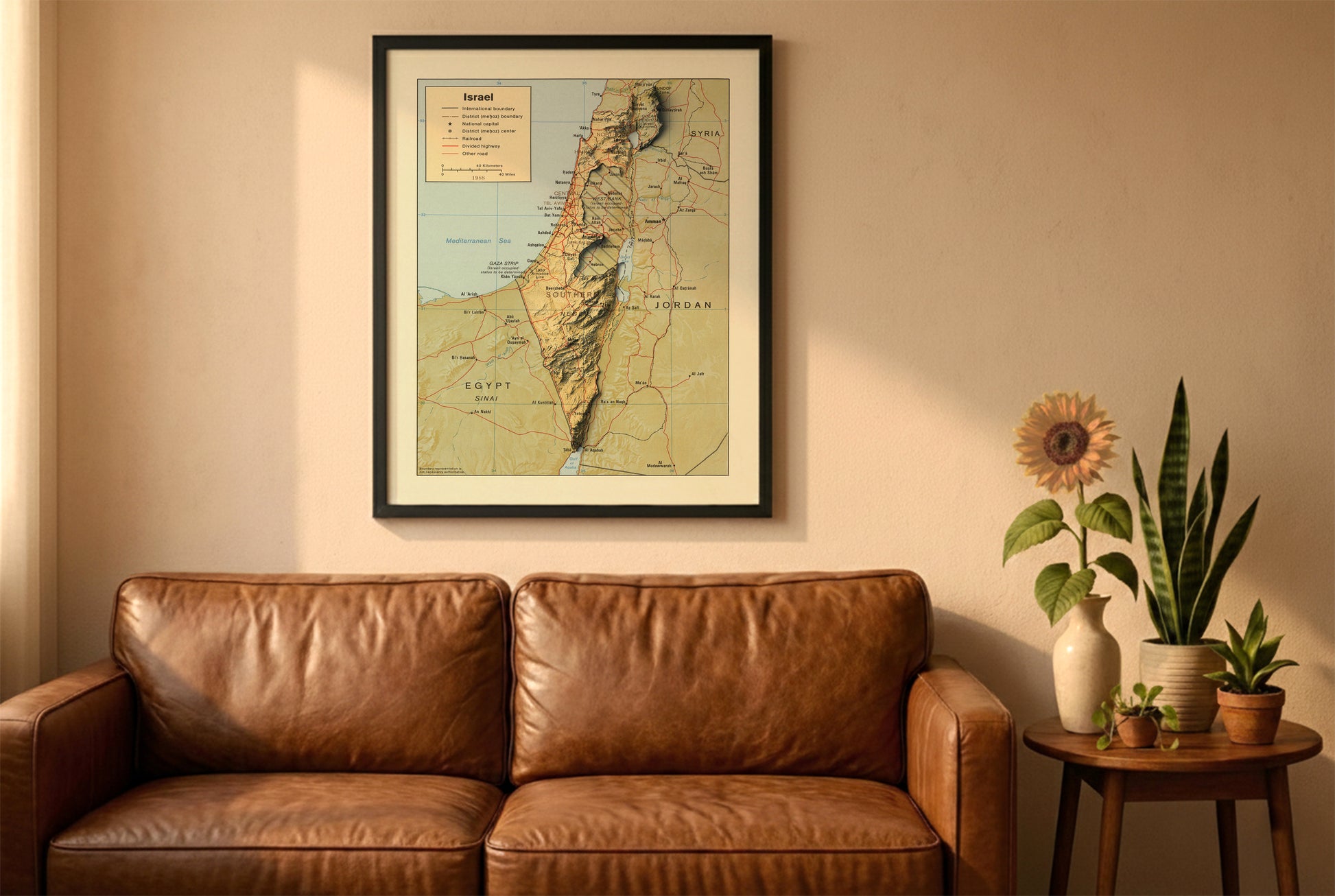 Israel vintage relief map in a black frame above a brown leather sofa. Boho living room with warm sunlight, sunflower, and indoor plants on a wooden side table