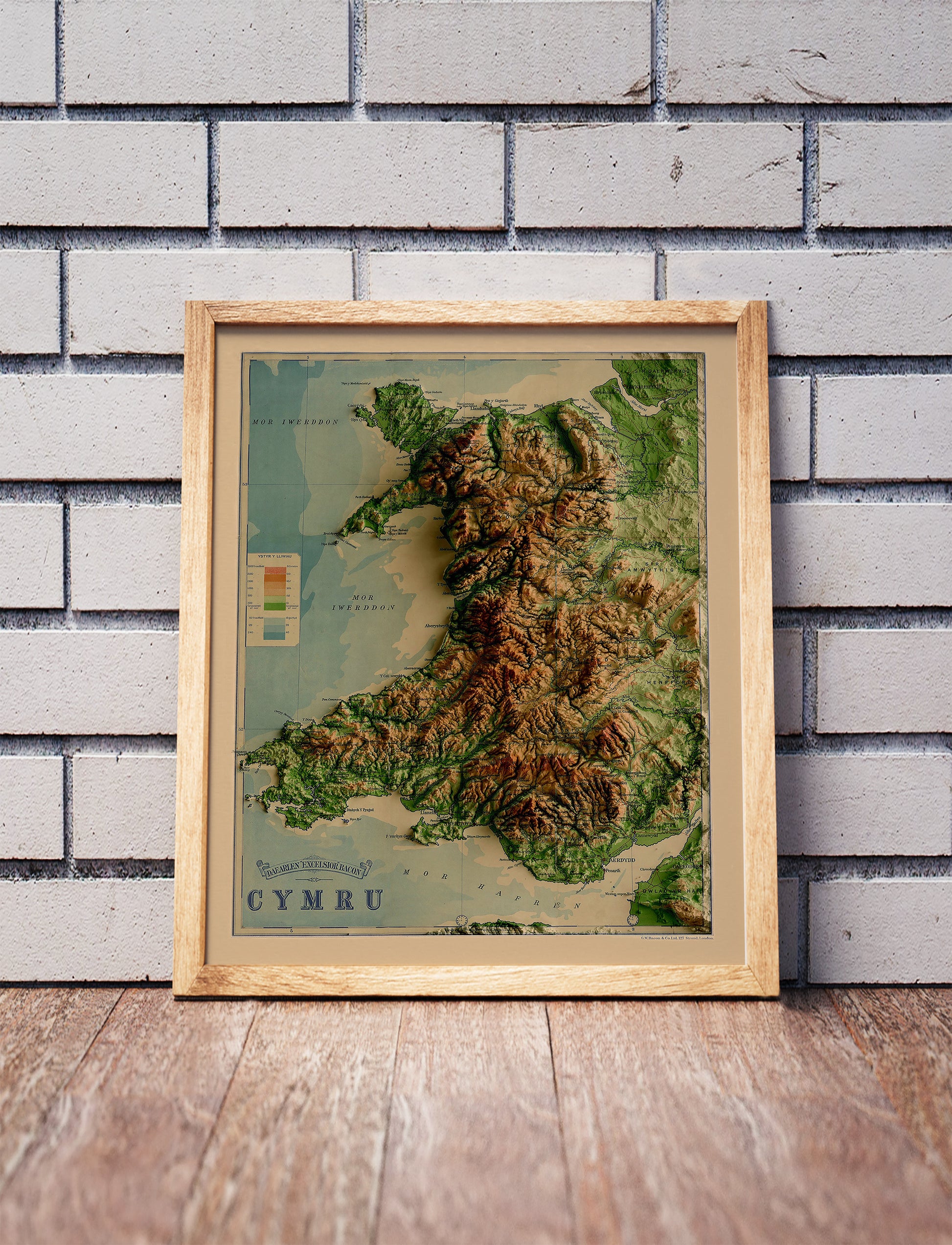 Vintage Wales shaded relief map in a wooden frame. Realistic terrain art print for home decor, displayed against a brick wall
