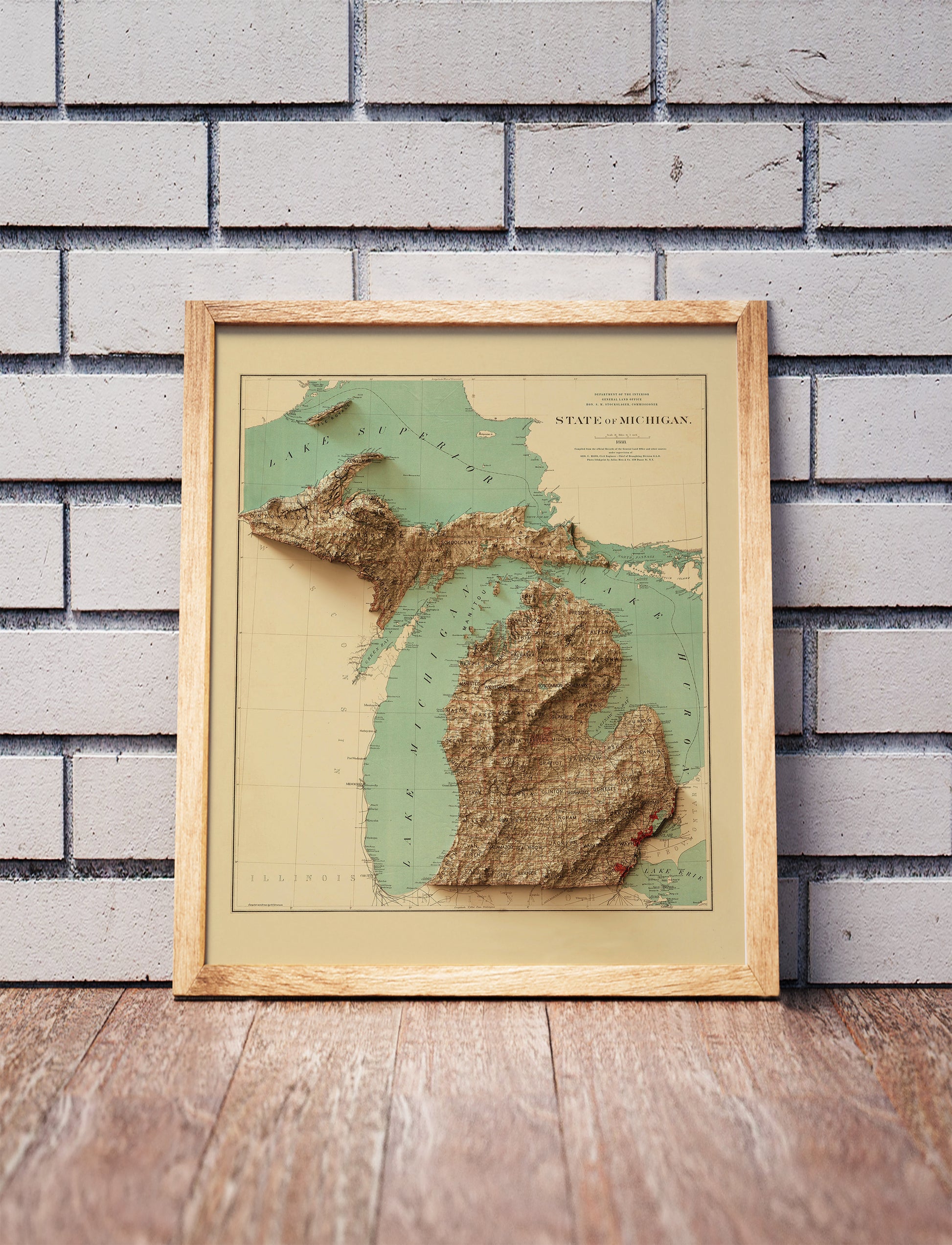 Vintage Michigan shaded relief map in a wooden frame. Realistic terrain art print for home decor, displayed against a brick wall