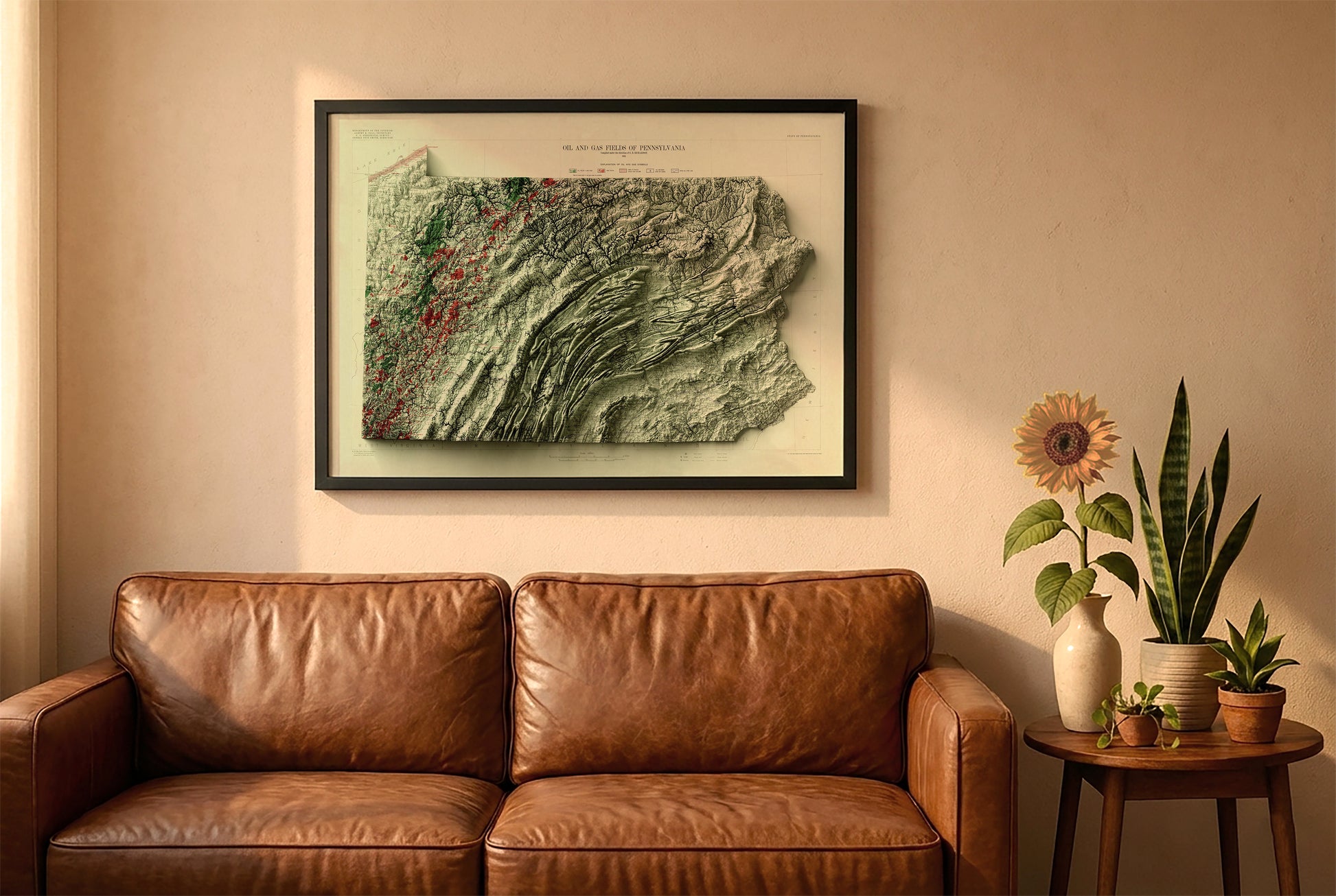 Pennsylvania vintage relief map in a black frame above a brown leather sofa. Boho living room with warm sunlight, sunflower, and indoor plants on a wooden side table