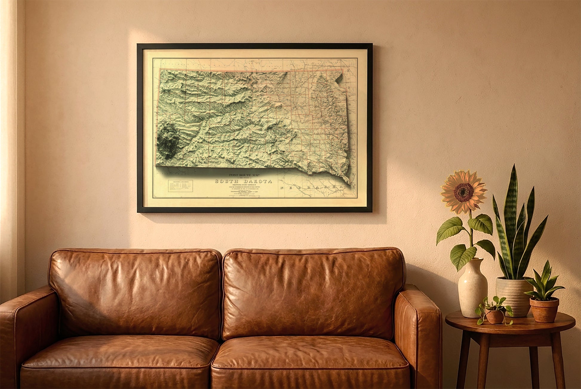 South Dakota vintage relief map in a black frame above a brown leather sofa. Boho living room with warm sunlight, sunflower, and indoor plants on a wooden side table