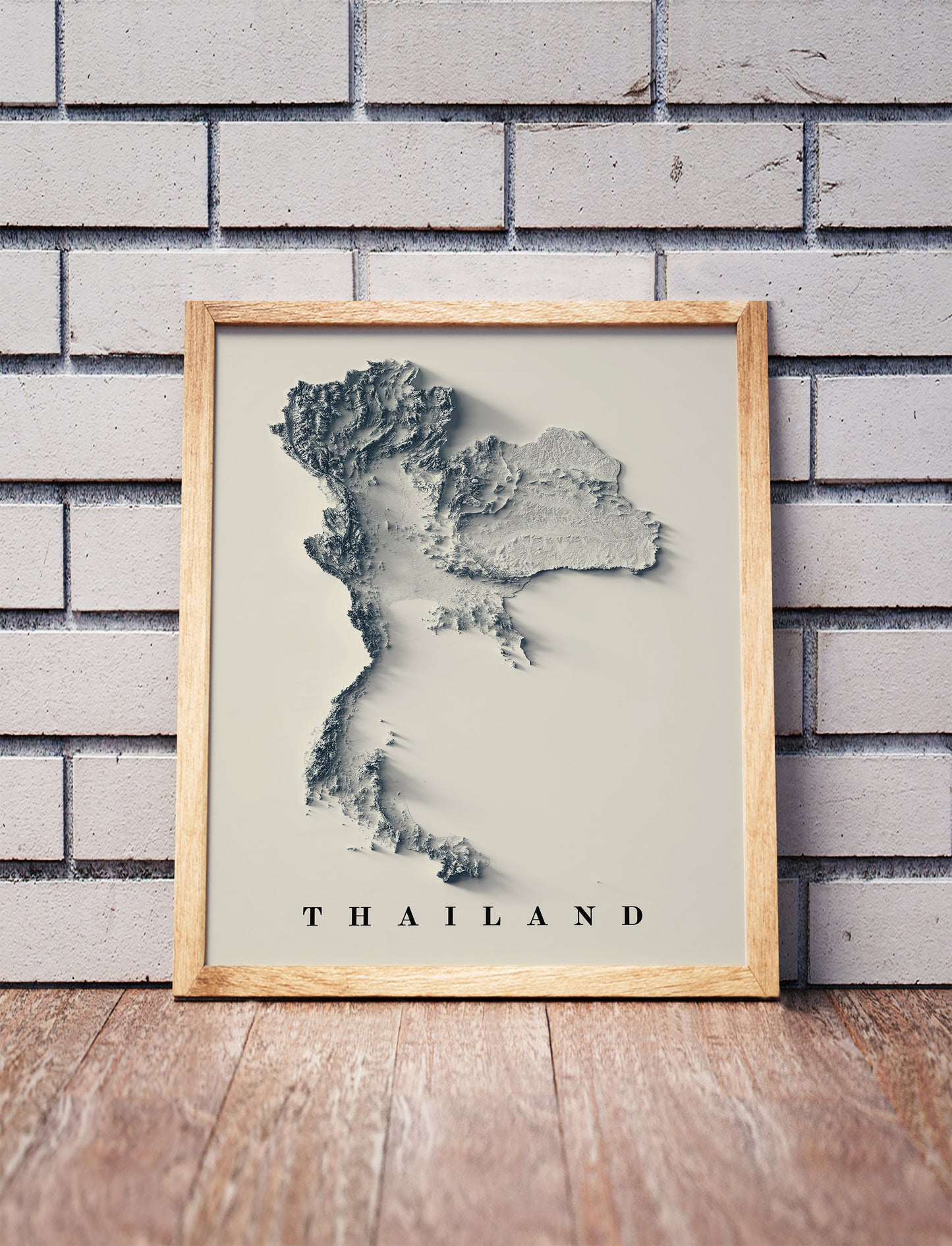 Thailand shaded relief map in a wooden frame. Realistic terrain art print for home decor, displayed against a brick wall