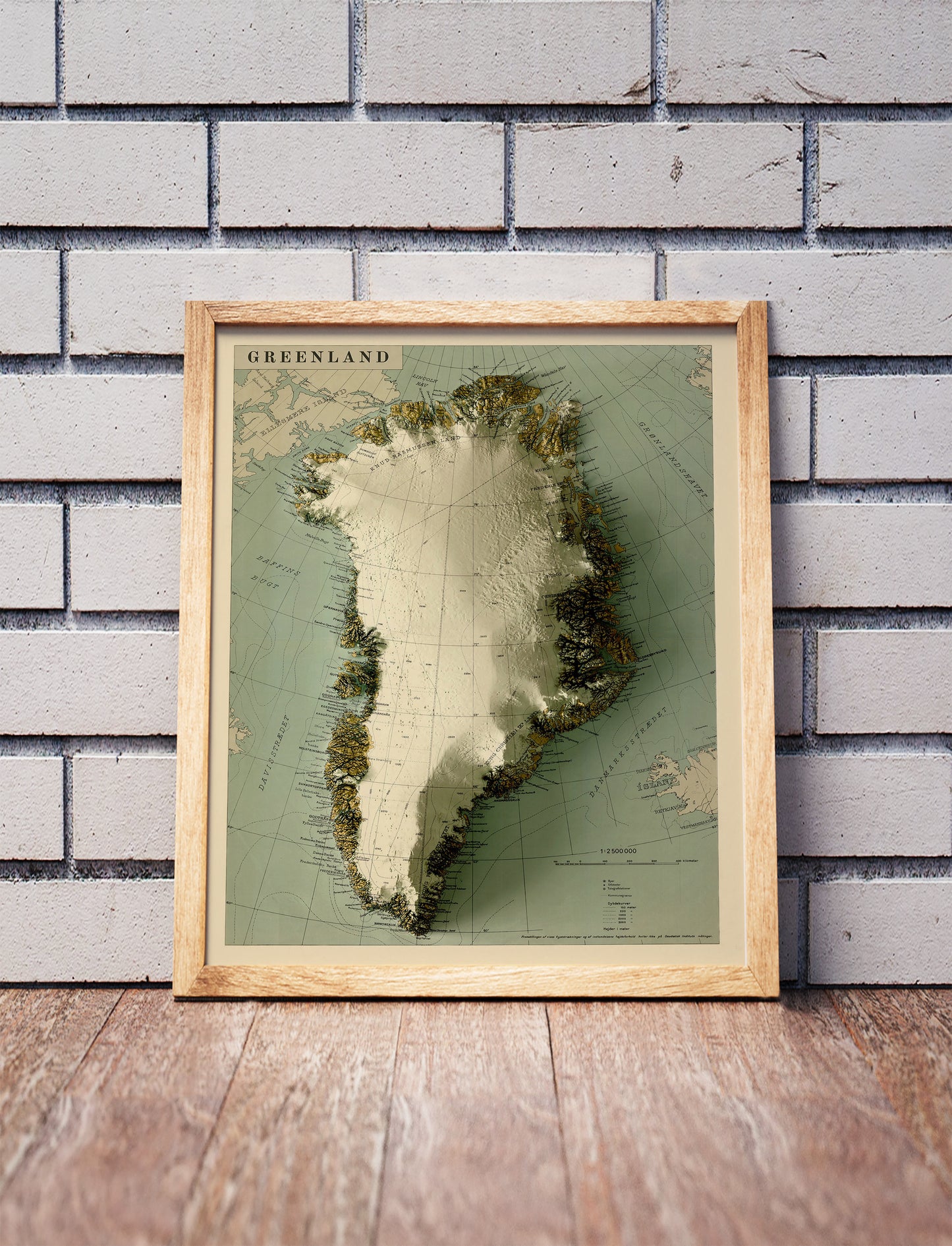 Vintage Greenland shaded relief map in a wooden frame. Realistic terrain art print for home decor, displayed against a brick wall