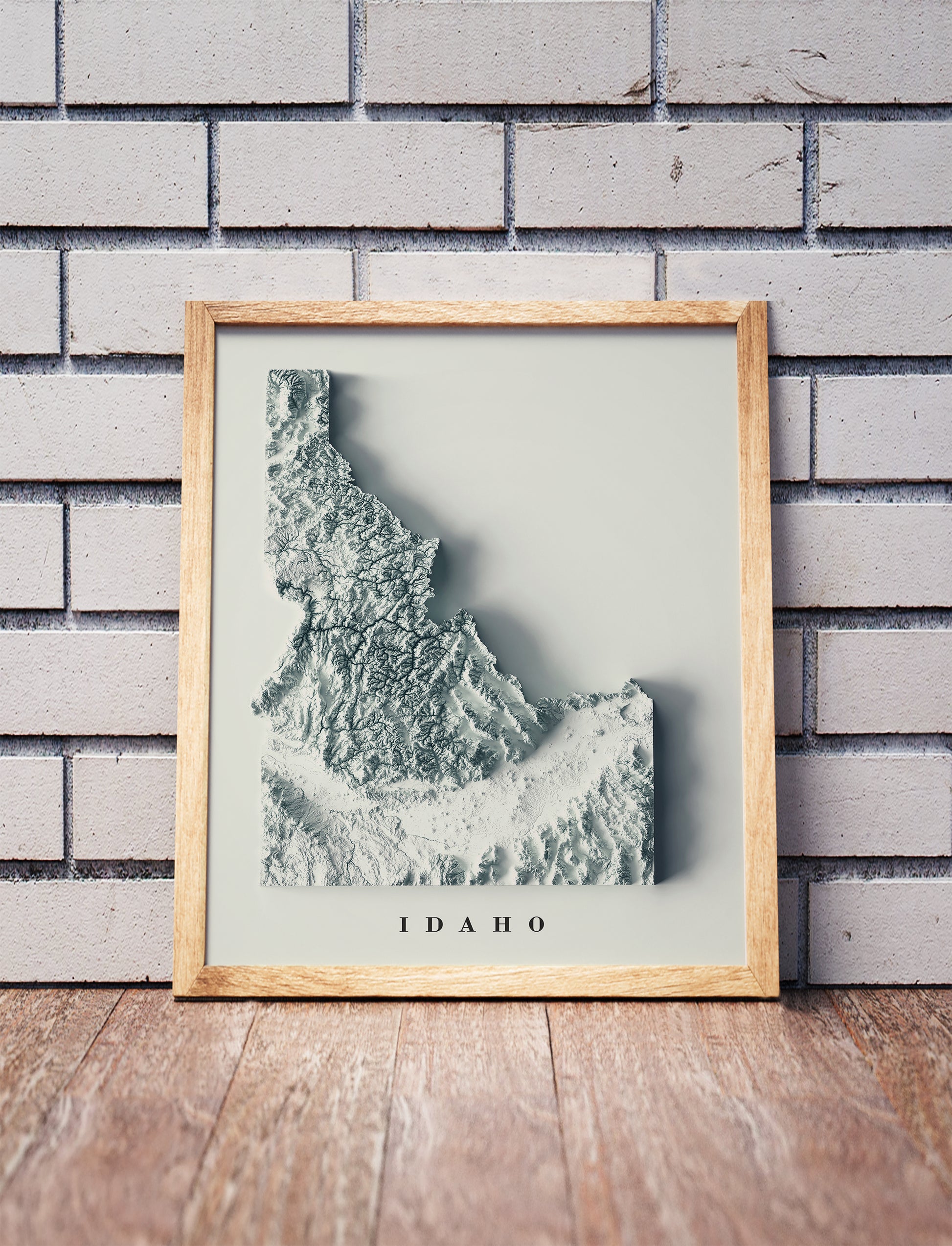 Idaho shaded relief map in a wooden frame. Realistic terrain art print for home decor, displayed against a brick wall