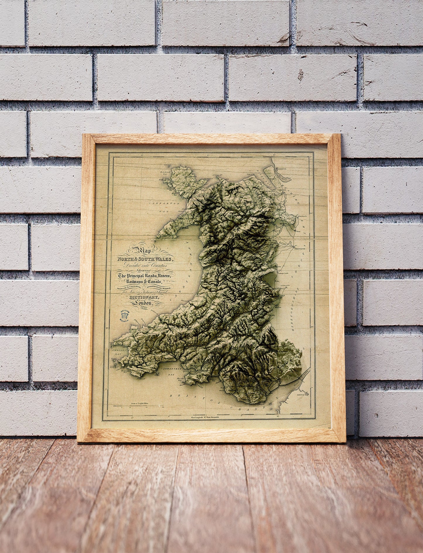 Vintage Wales shaded relief map in a wooden frame. Realistic terrain art print for home decor, displayed against a brick wall