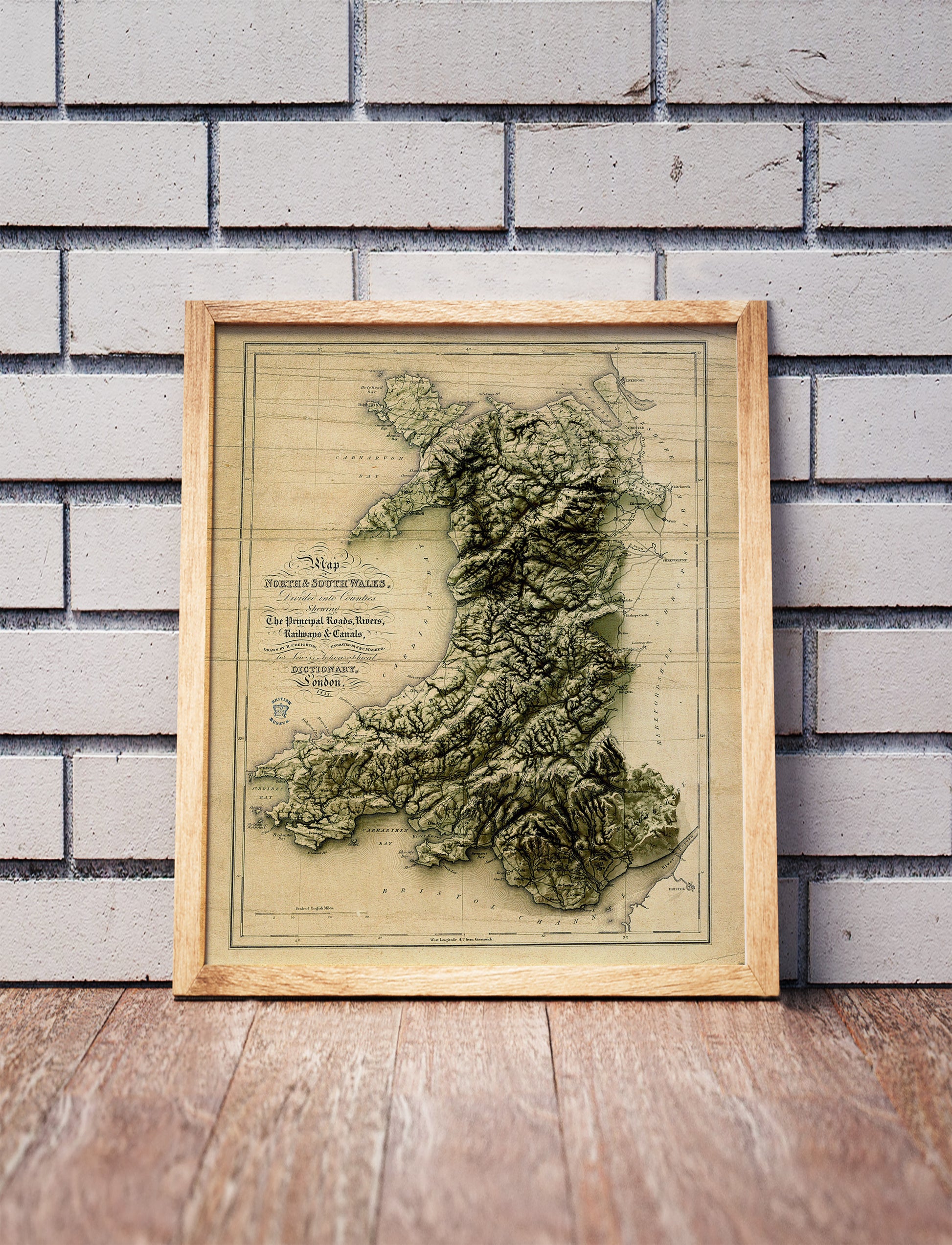 Vintage Wales shaded relief map in a wooden frame. Realistic terrain art print for home decor, displayed against a brick wall