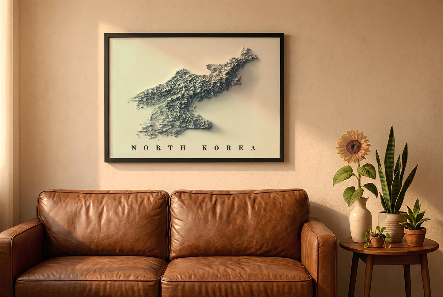 North Korea relief map in a black frame above a brown leather sofa. Boho living room with warm sunlight, sunflower, and indoor plants on a wooden side table